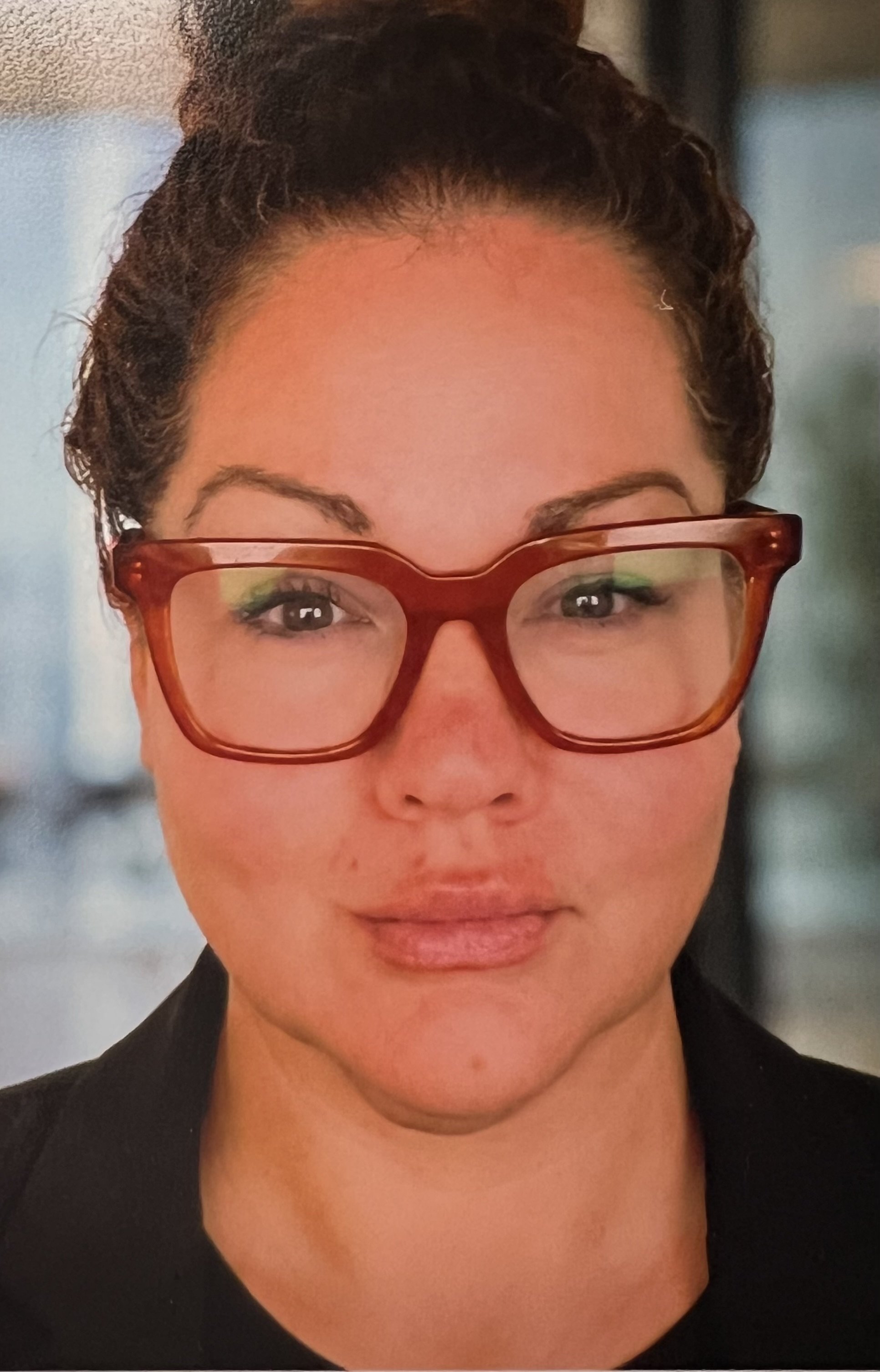 A woman with dark hair styled in a bun, wearing red glasses, and a black top, with a calm expression. a soul lead person, a soul coach.