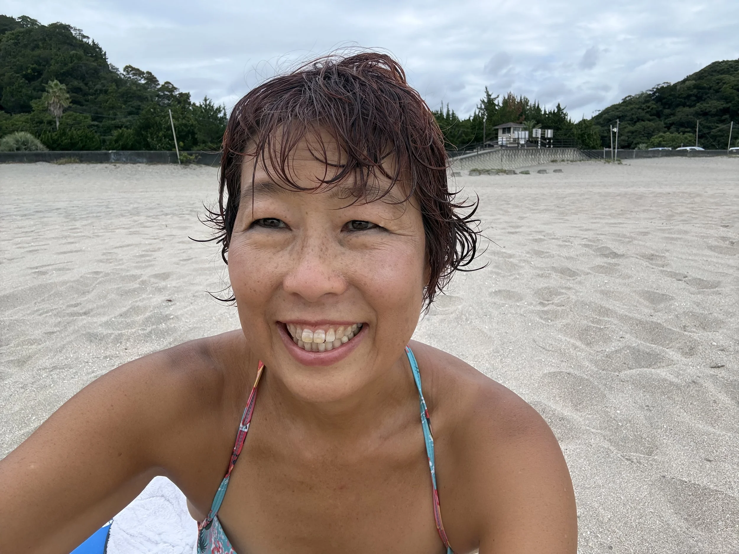 ビーチで笑顔の女性の顔の一部が写った写真。背景には山と木々、空が見える。