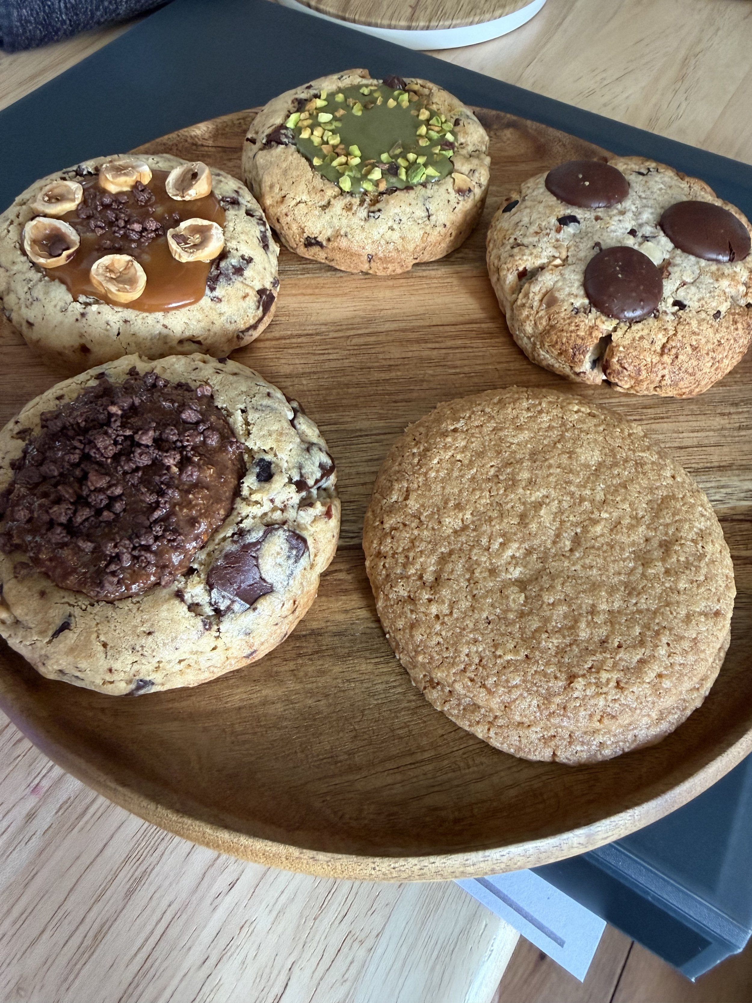 木製の皿に乗った五つのクッキー。上から順にキャラメルとナッツ、抹茶とチョコレート、チョコレートとナッツ、チョコレートとクッキークランチ、シンプルなオートミールクッキー。