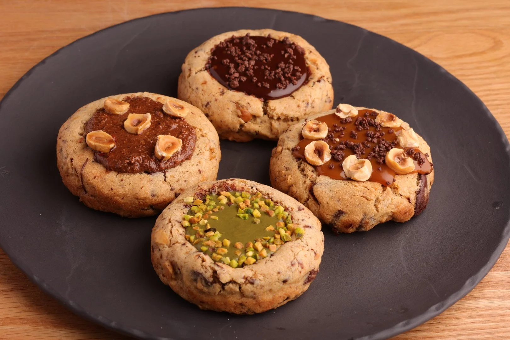 4つのクッキーが黒いプレートに乗っている。各クッキーは異なるトッピングが施されている。1つはピスタチオとウイナーストリーの組み合わせ、他はチョコレートとナッツなどのトッピングがされている。