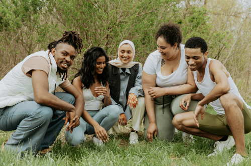 Five diverse young adults crouching and smiling on grass outdoors in a park.