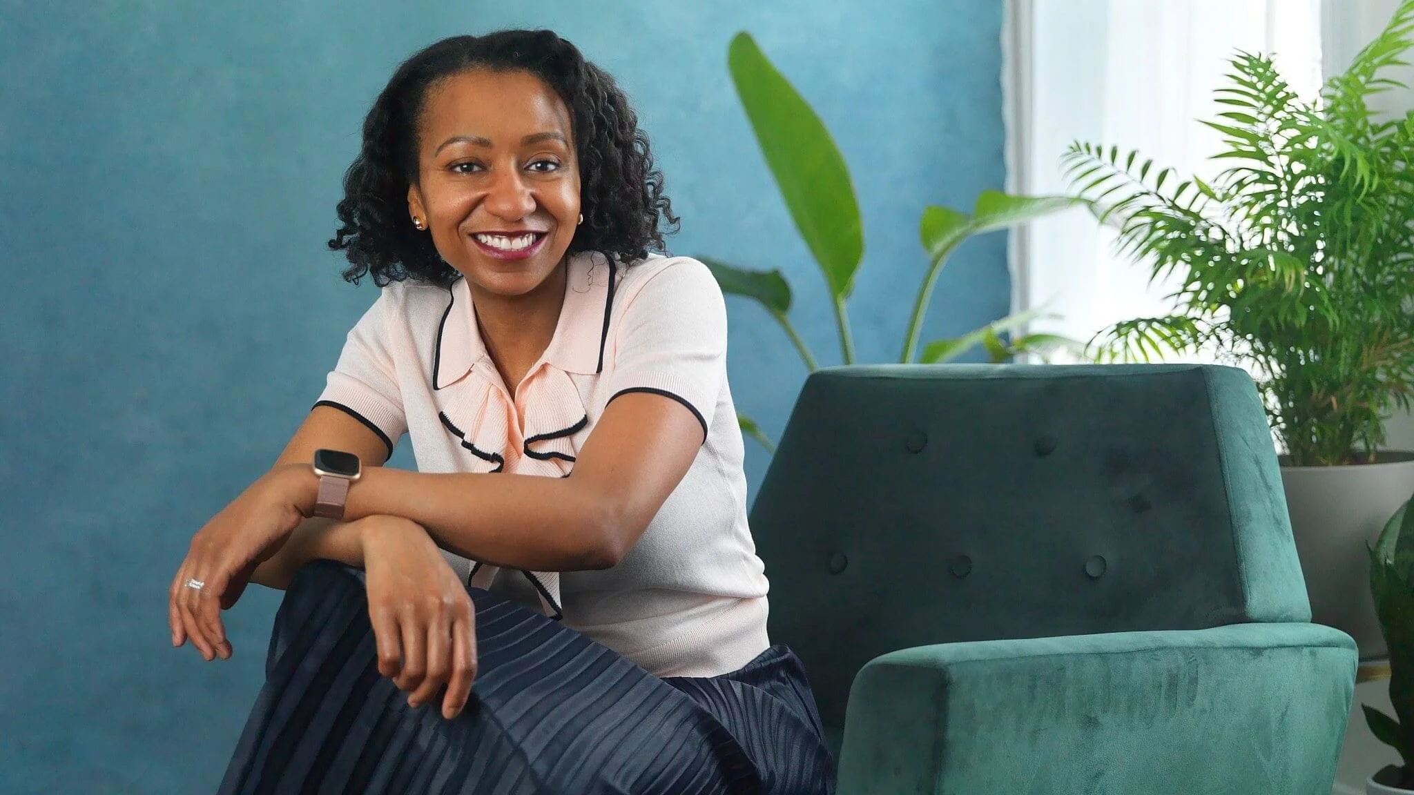 A smiling woman with curly black hair, wearing a light pink shirt with black accents, sitting on a teal upholstered chair in a room with a blue wall and green plants.