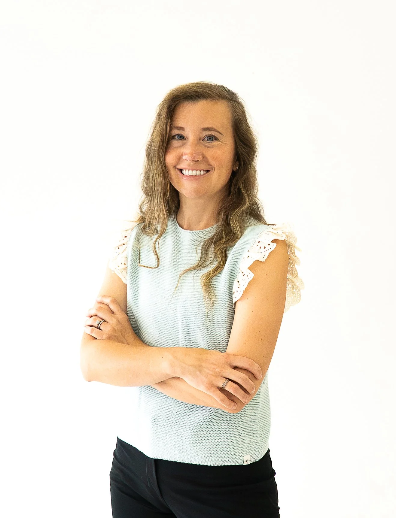 A woman with long wavy brown hair, wearing a cream-colored top with lace details on the sleeves, smiling with arms crossed, standing against a white background.