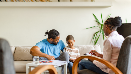 A man, a young boy, and a woman are sitting on a beige couch in a living room, engaging in a conversation or activity together.