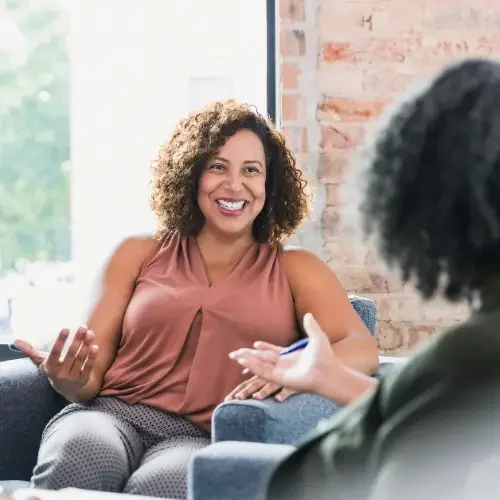 A woman with curly hair smiling and talking to another person in a therapy or counseling session, sitting on a blue couch near a window with a brick wall in the background.
