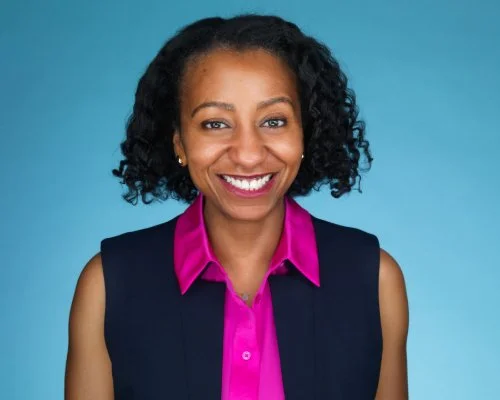 A woman with curly black hair smiling, wearing a bright pink shirt and a dark vest, against a blue background.