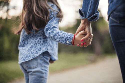 A child holding an adult's hand outdoors, with trees and a path in the background.
