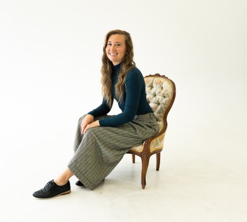 A woman with long wavy hair, wearing a blue long sleeve shirt, plaid skirt, and black shoes, smiling and sitting sideways on an ornate vintage armchair with a wooden frame and cushioned back, against a plain white background.