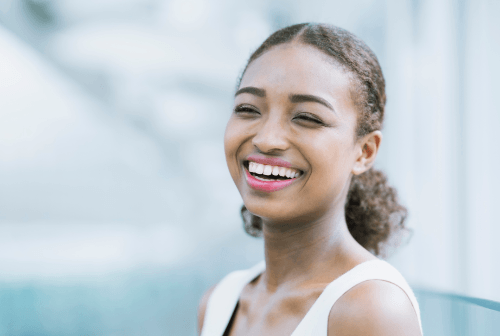 A smiling woman with curly hair in a white sleeveless top, sitting near a window.