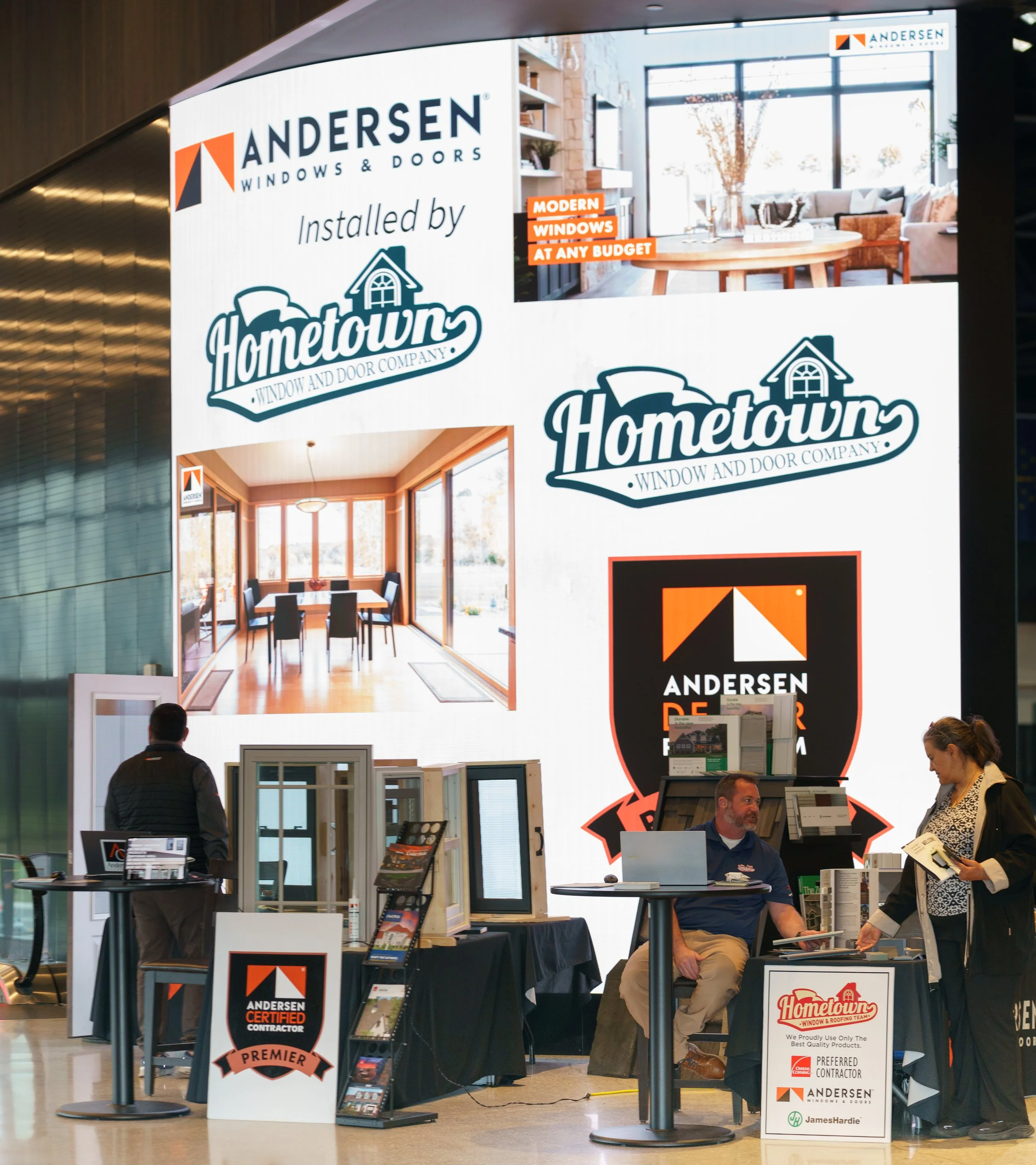 Booth for Andersen Windows & Doors at an indoor trade show, featuring promotional materials, displays of window and door samples, and representatives interacting with visitors.