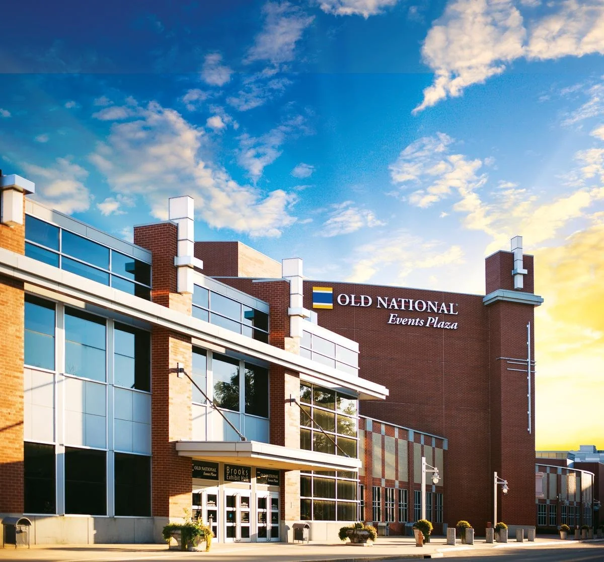 Exterior view of Old National Events Plaza building at sunset, with brick walls, large glass windows, and signage, under a partly cloudy sky.