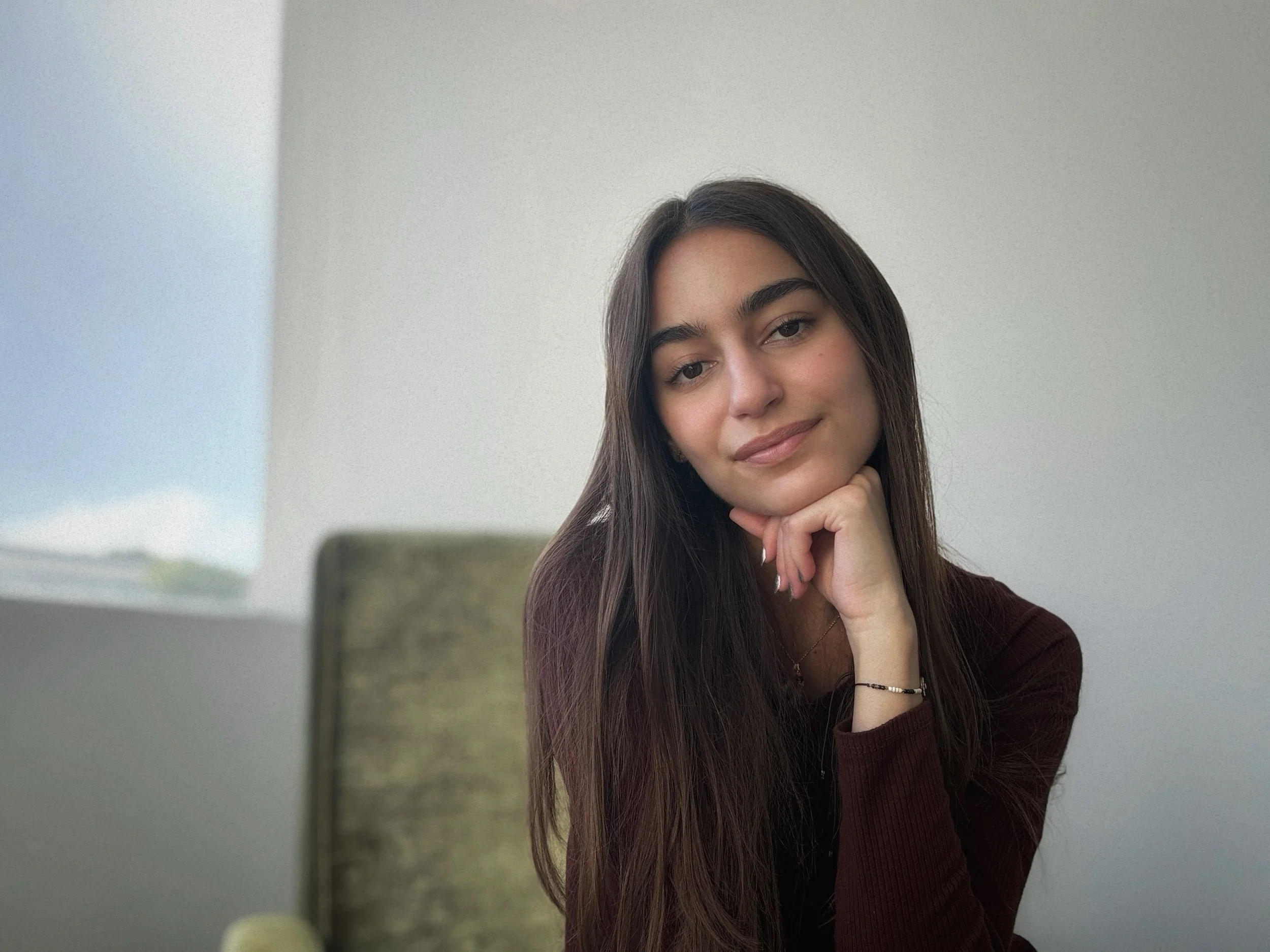 Close-up of a young woman with long brown hair, lightly smiling, resting her chin on her hand, in a room with soft natural lighting and neutral-colored walls.