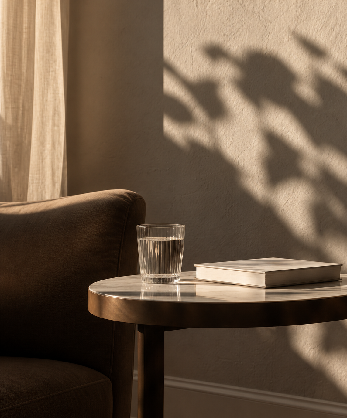 A cozy corner with a brown armchair, a round marble-top table with a glass of water, a closed book, and sunlight casting shadows of leaves on the wall.