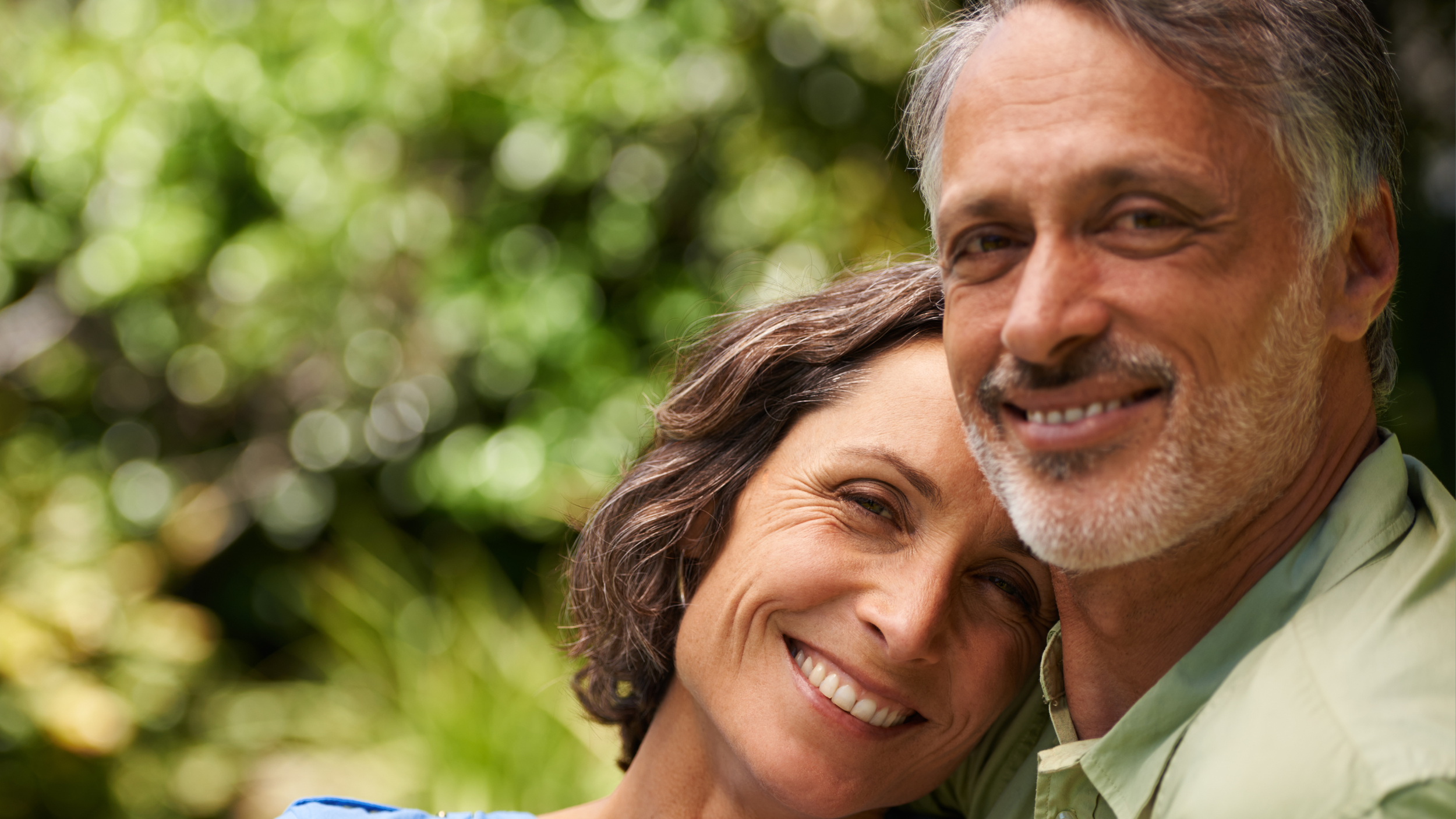 Close-up of an elderly couple smiling and hugging outdoors with green bokeh background.