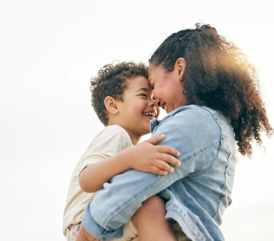 A woman holding a young boy, both smiling with their faces close together against a white background.