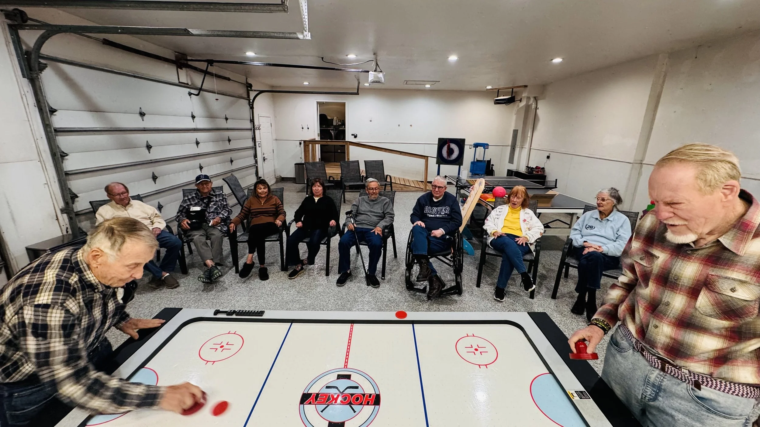 A group of elderly people sitting in chairs in a garage, playing air hockey and watching the game, with a man in the foreground holding a hockey paddle.