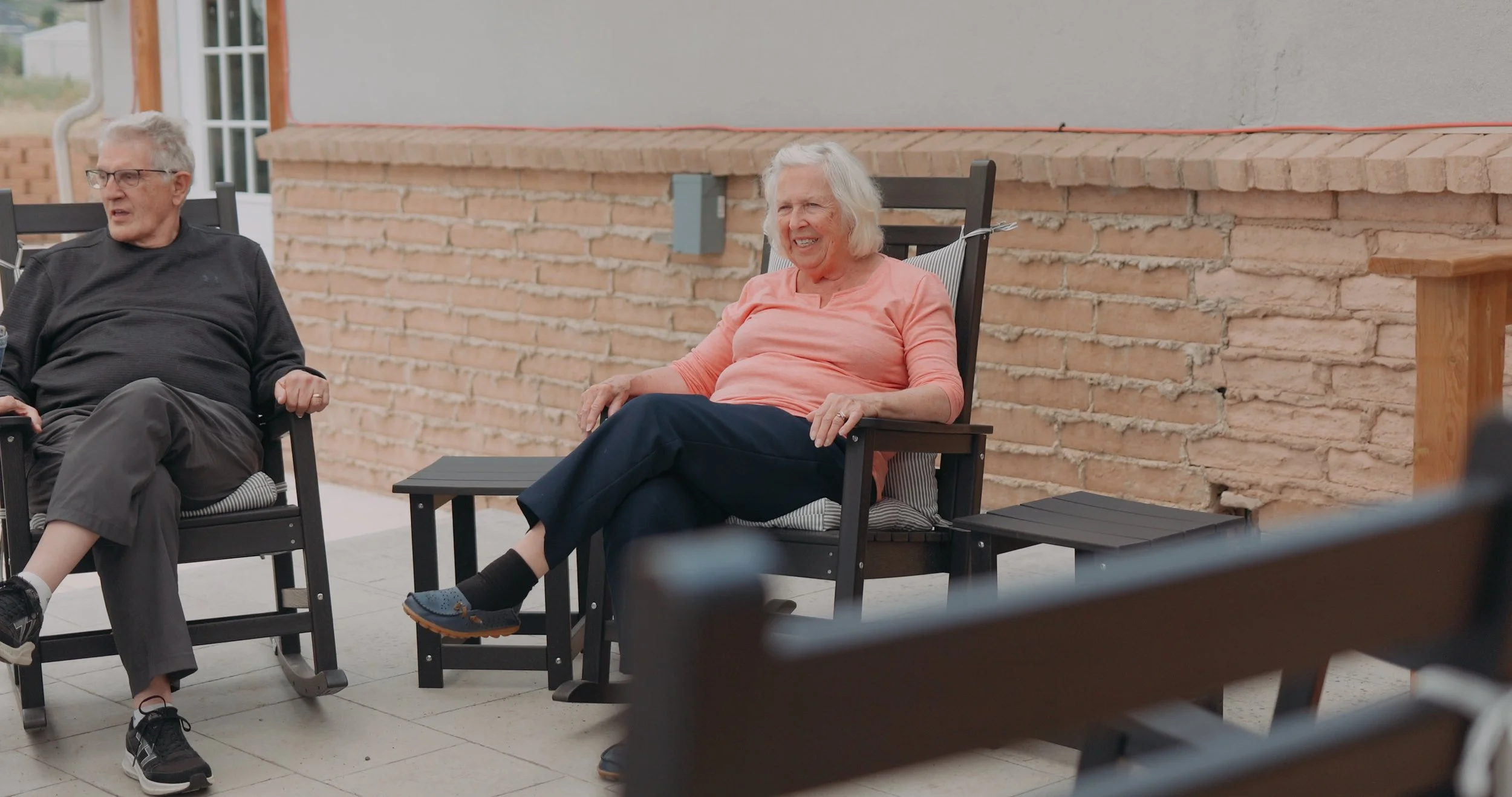 Senior man and woman sitting outside on patio chairs, smiling and talking, with brick wall background.