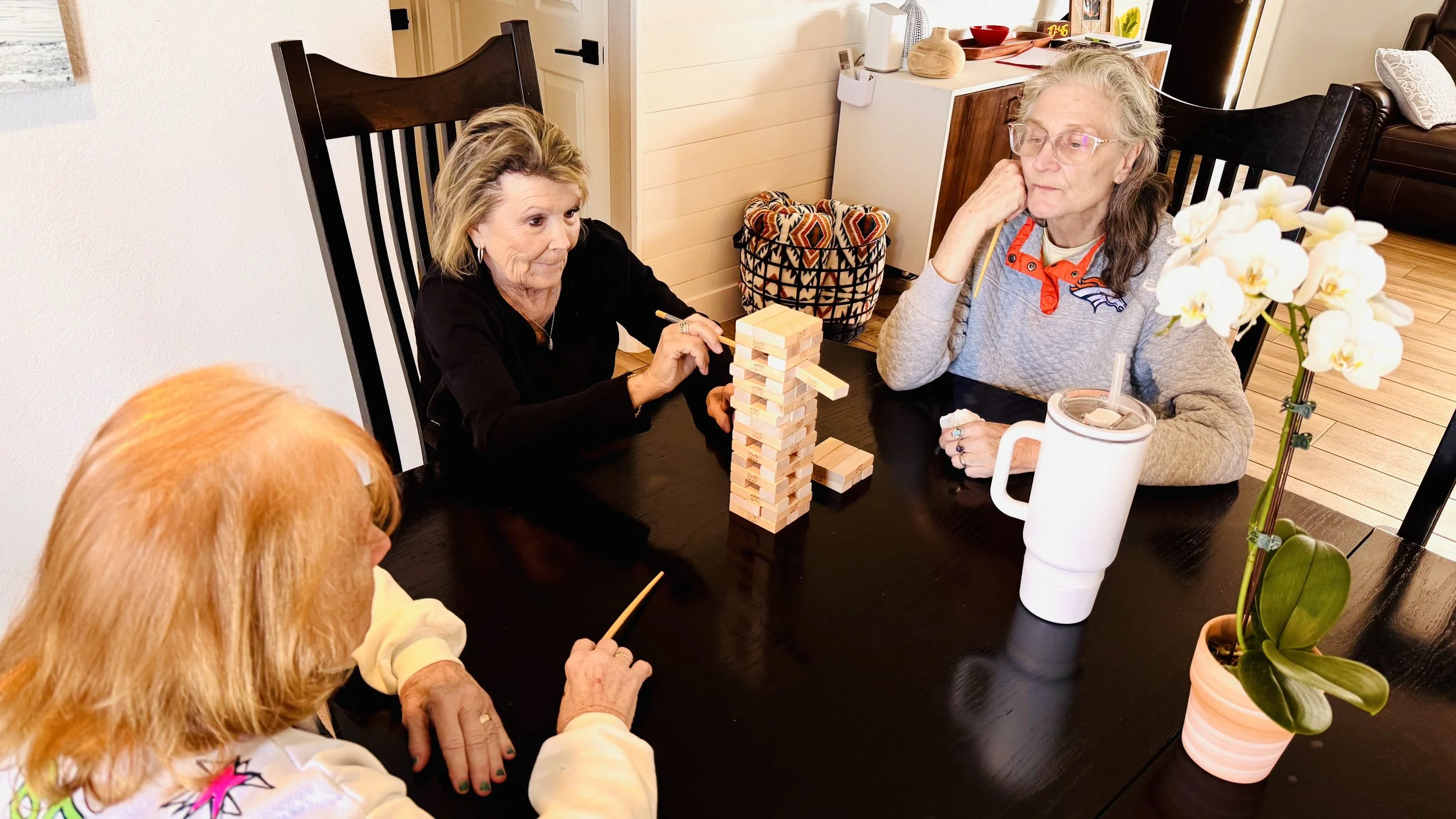 Four elderly women playing a game of Jenga at a dining table, with a potted orchid flower and a mug on the table.
