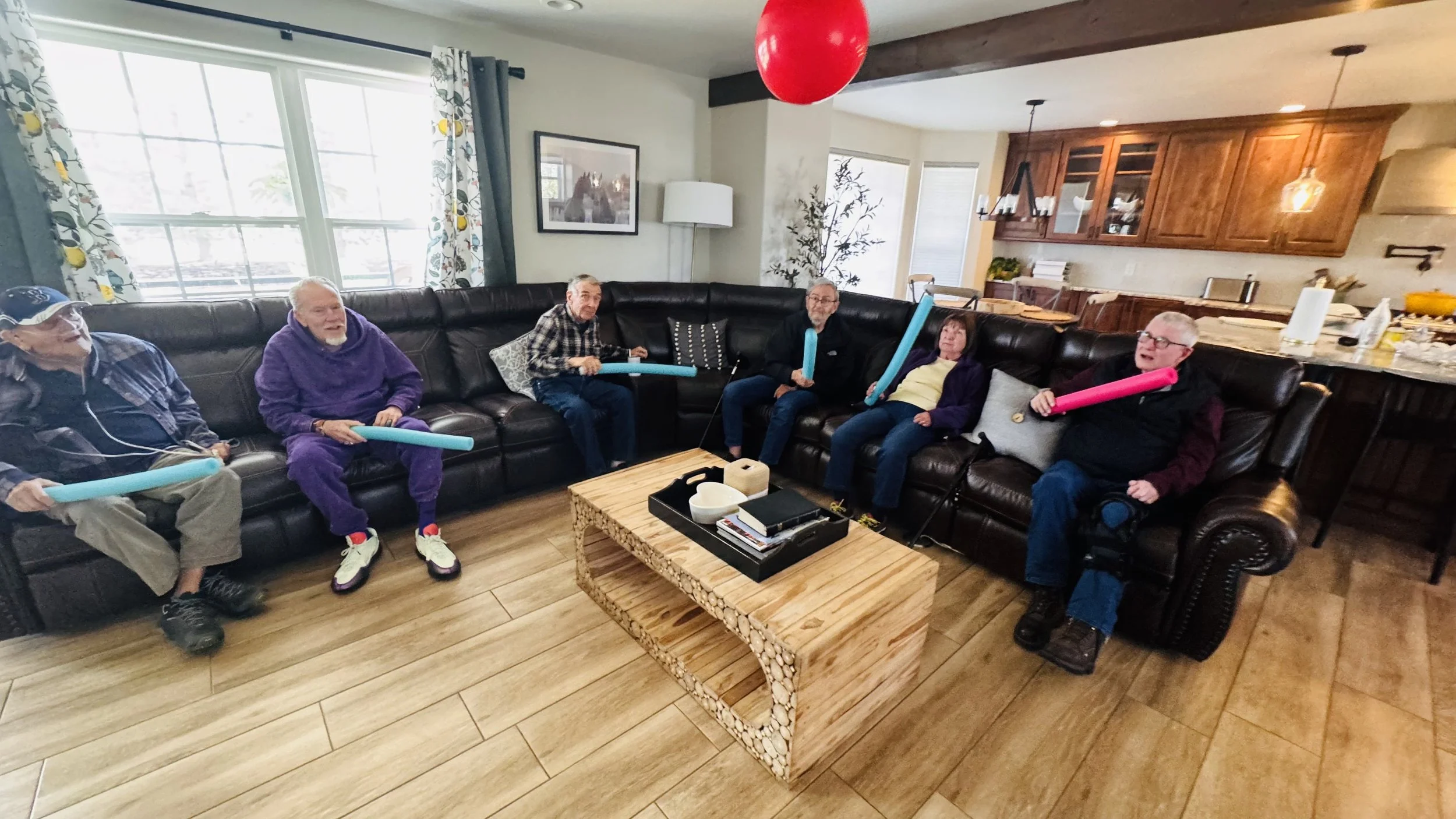 Six elderly people sitting on a black leather couch in a living room, holding colorful pool noodles. The room has large windows with floral curtains, a wooden coffee table with books and bowls, and a kitchen area in the background.