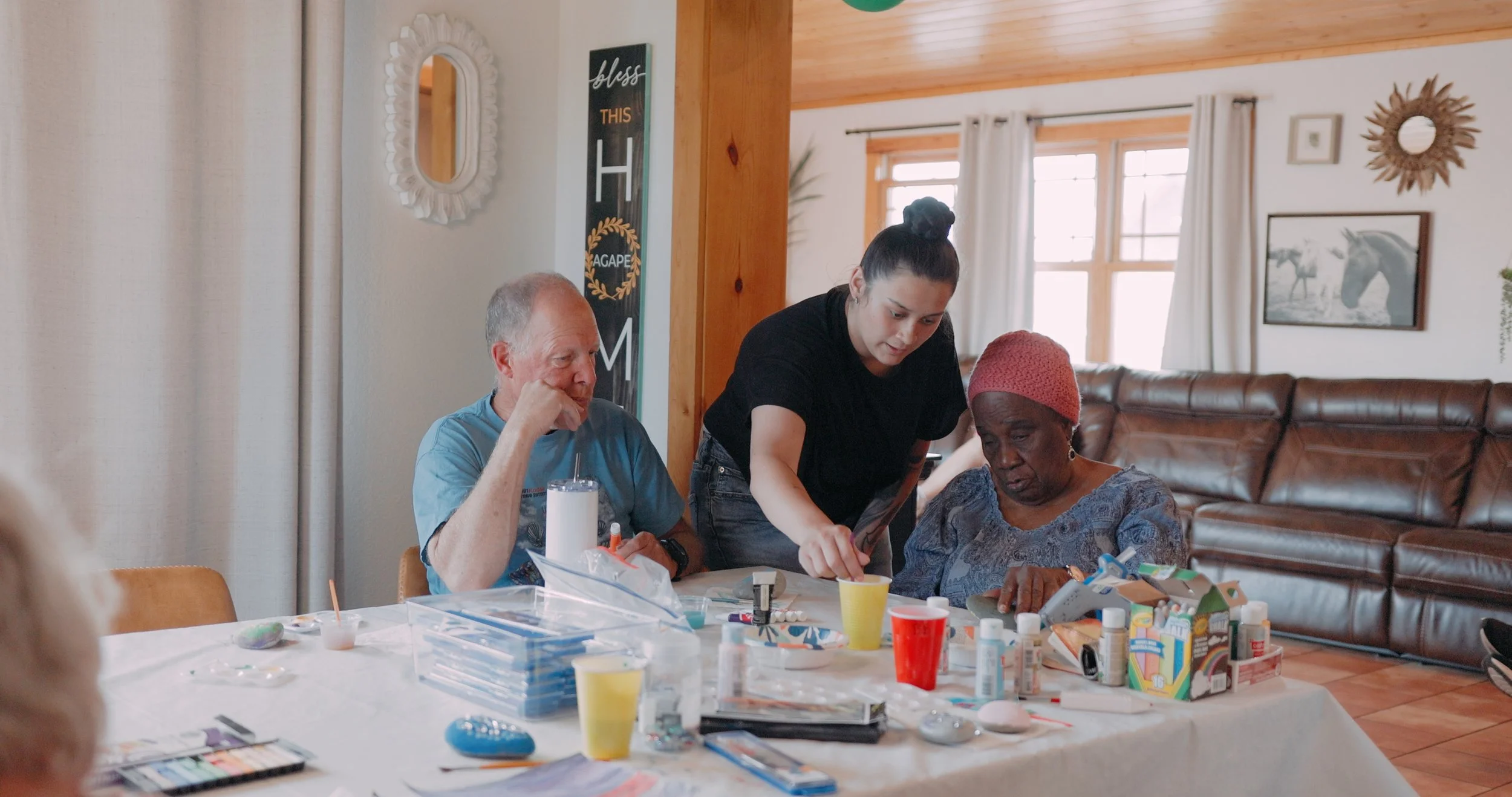 A young woman helping two elderly women with arts and crafts projects at a table in a living room.