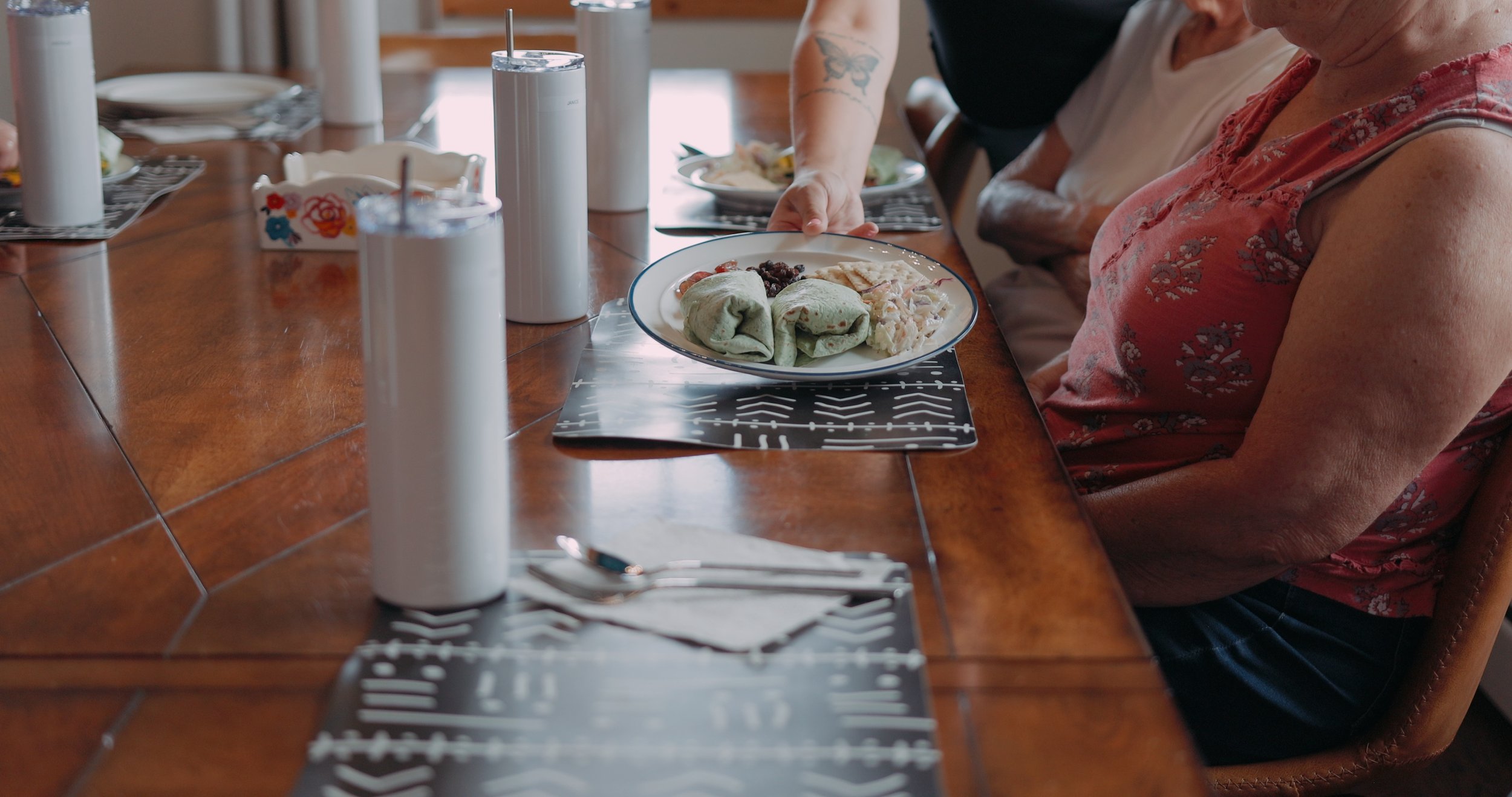 A person is serving food on a plate at a wooden dining table. The plate contains three items wrapped in green tortillas, some beans, and coleslaw. There are drinks in white cups with straws, and napkins with utensils on the table.