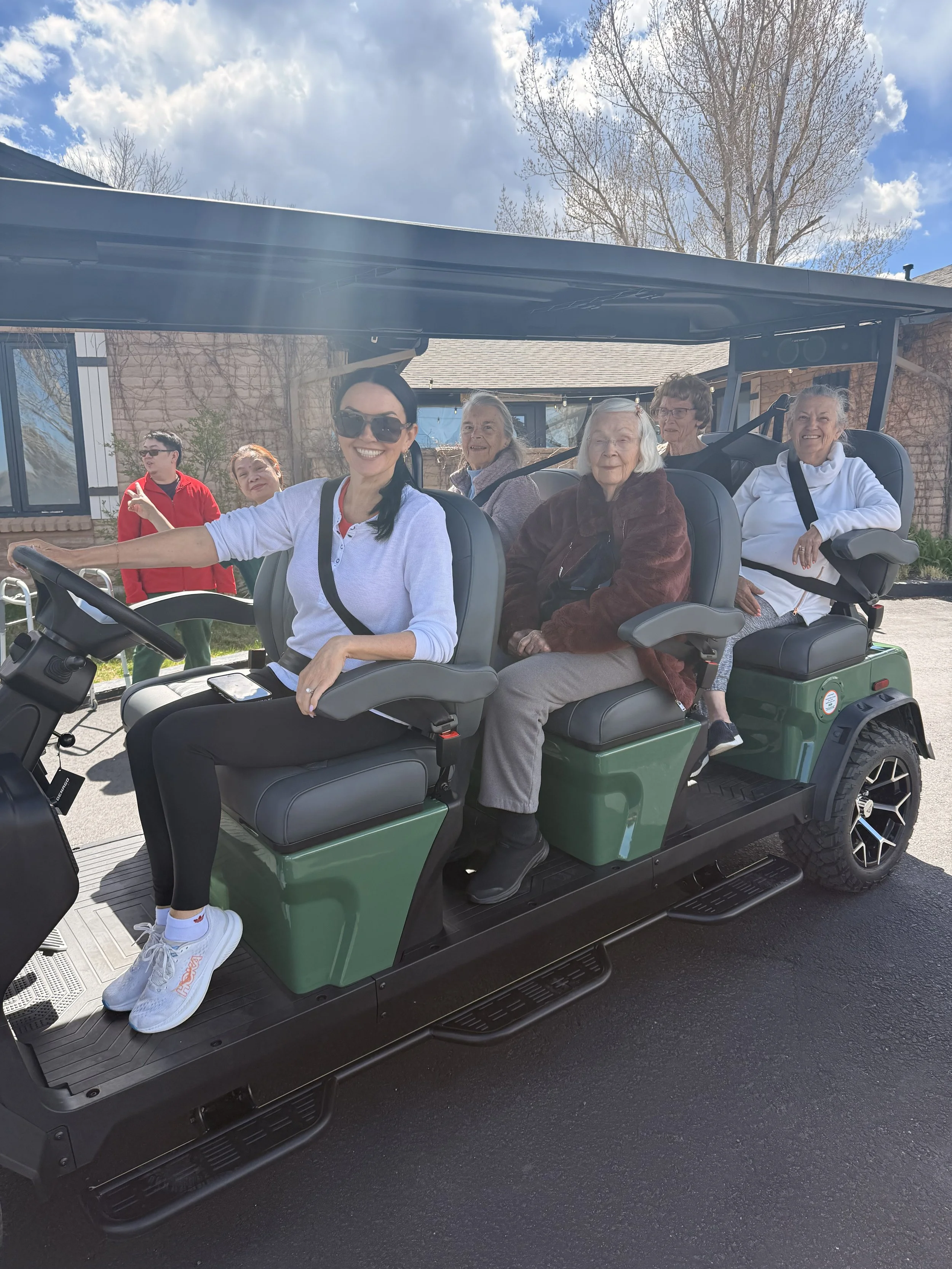 Six people, mostly elderly women, seated in a green electric golf cart. The cart is outdoors on a sunny day with a blue sky and some clouds. The background shows a building with brick walls and leafless trees.