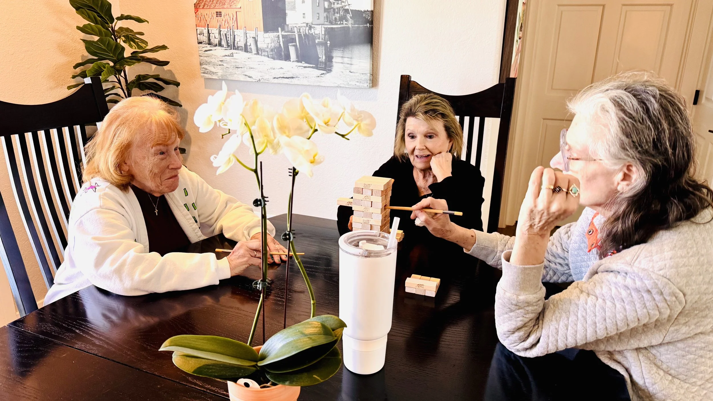 Four elderly women sitting around a dark wooden table playing a game of Jenga.