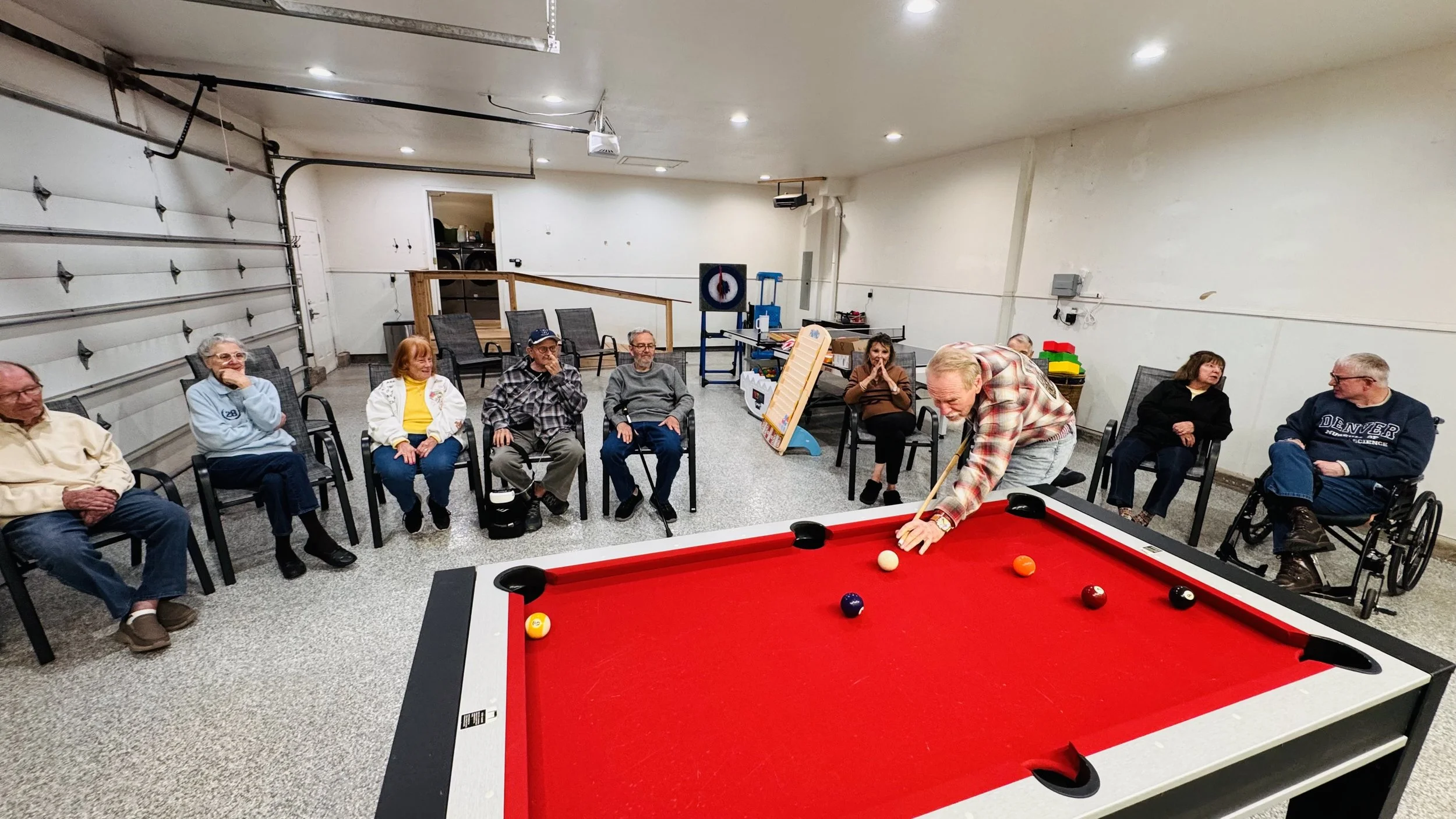 People sitting around a pool table in a recreation room, watching a man in a plaid shirt playing pool. The room has a white wall, some chairs, and exercise equipment in the background.