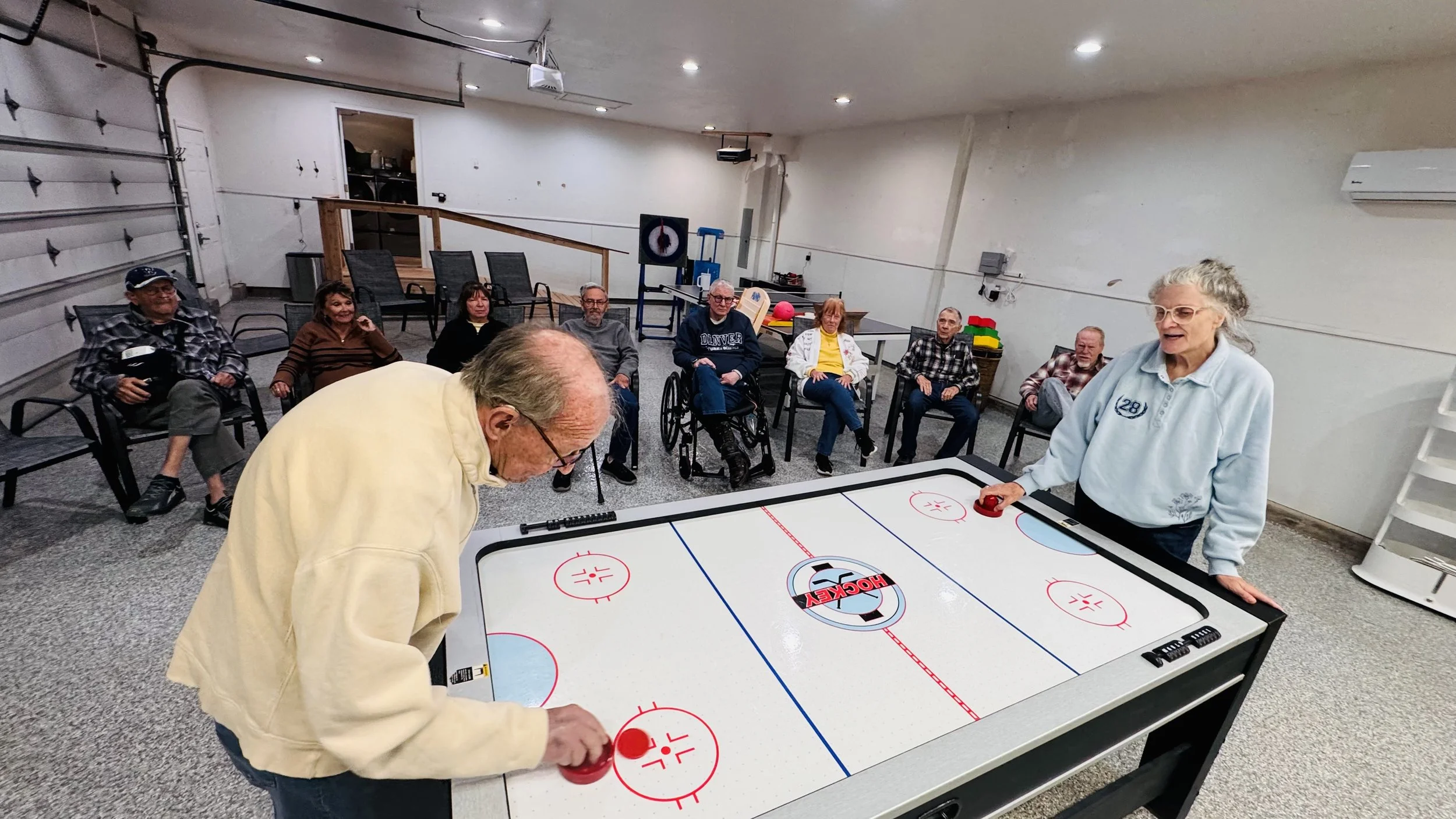 Group of elderly people sitting and watching as two women play air hockey on an air hockey table in a recreational room.