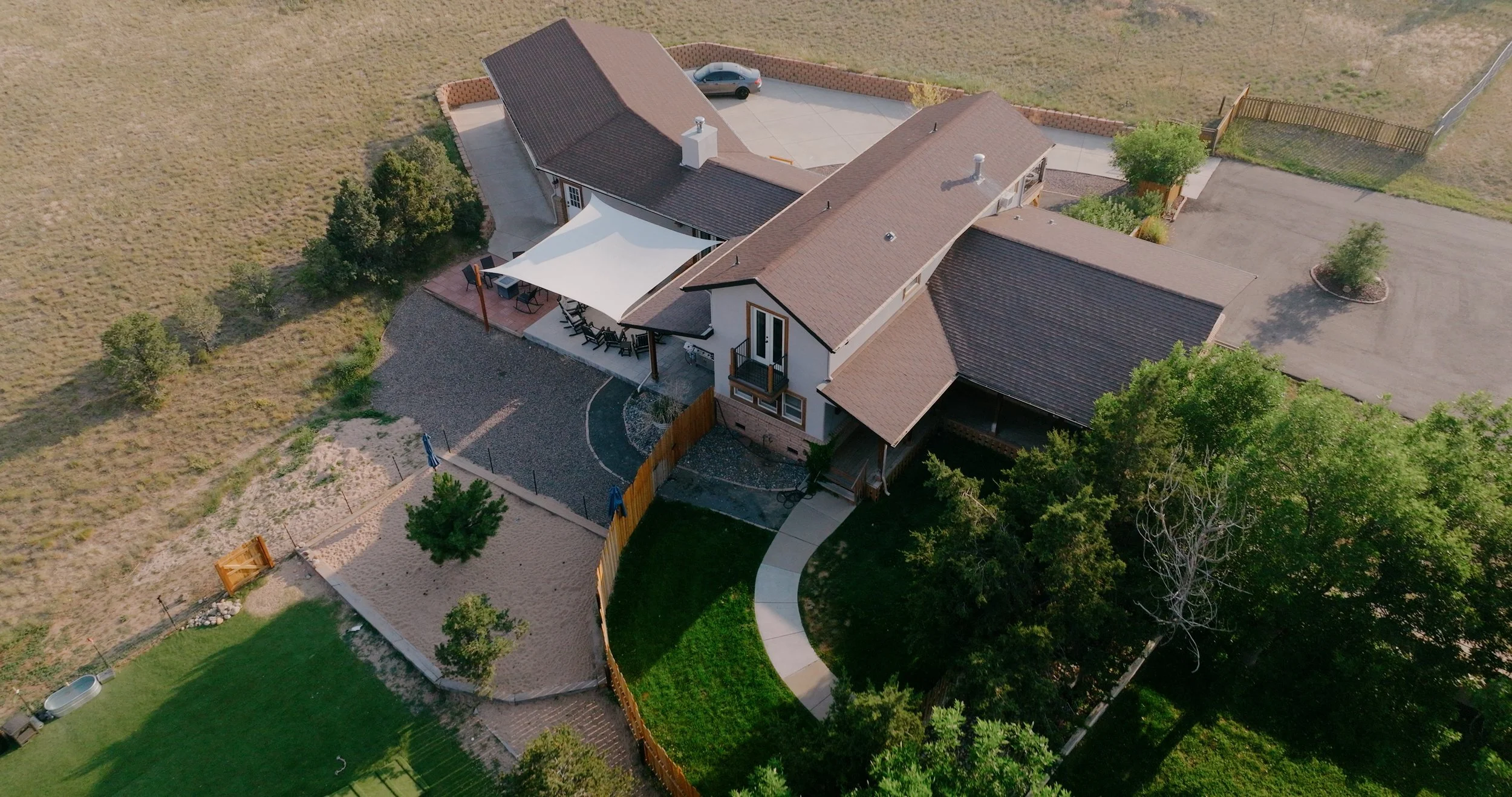 An aerial view of a house with a backyard, patio area with seating, a driveway with parked car, and surrounding trees and open fields.