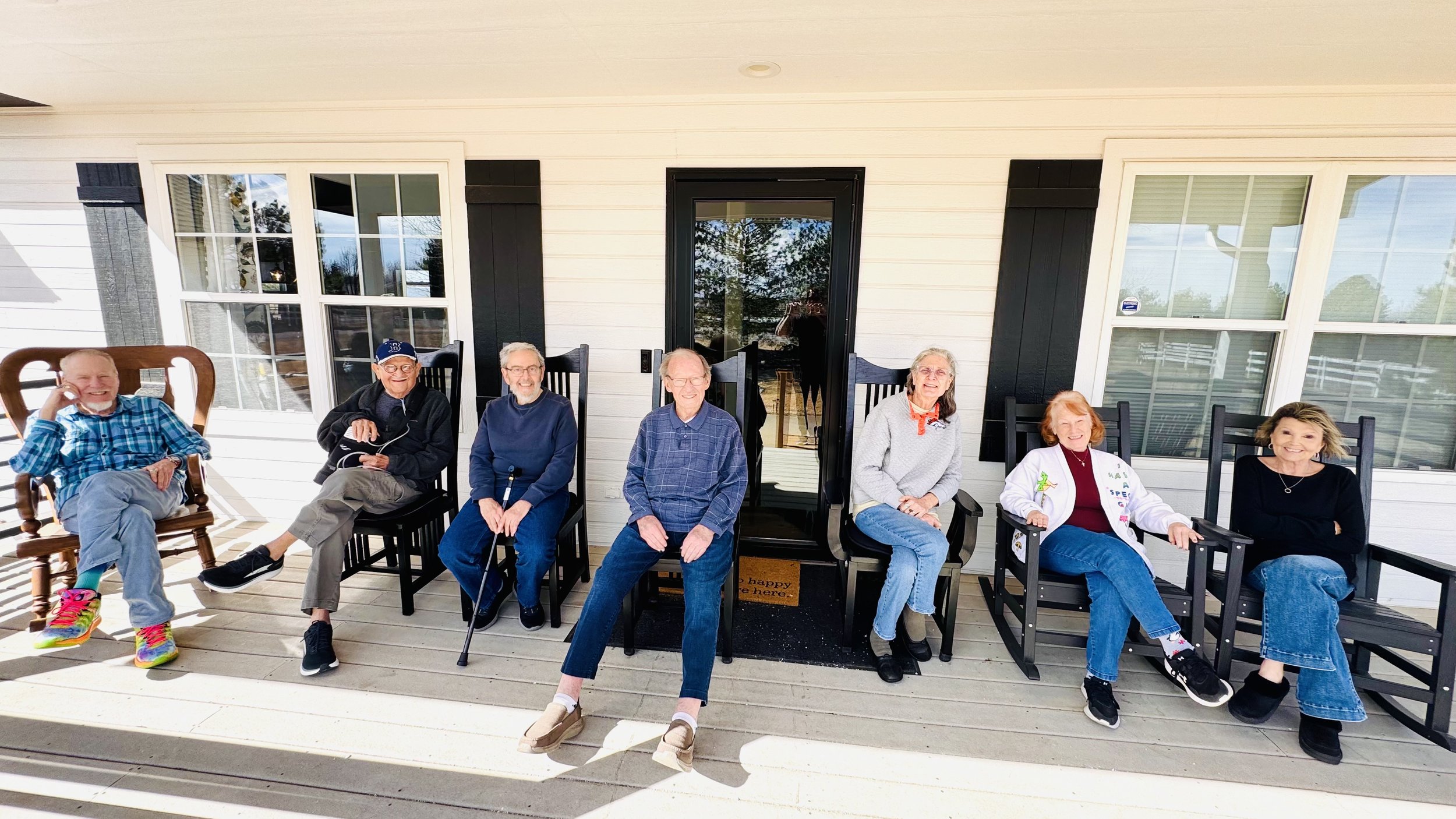 Group of seven seniors sitting on a porch with white siding house, large windows, black trim, and a glass door, enjoying a sunny day.