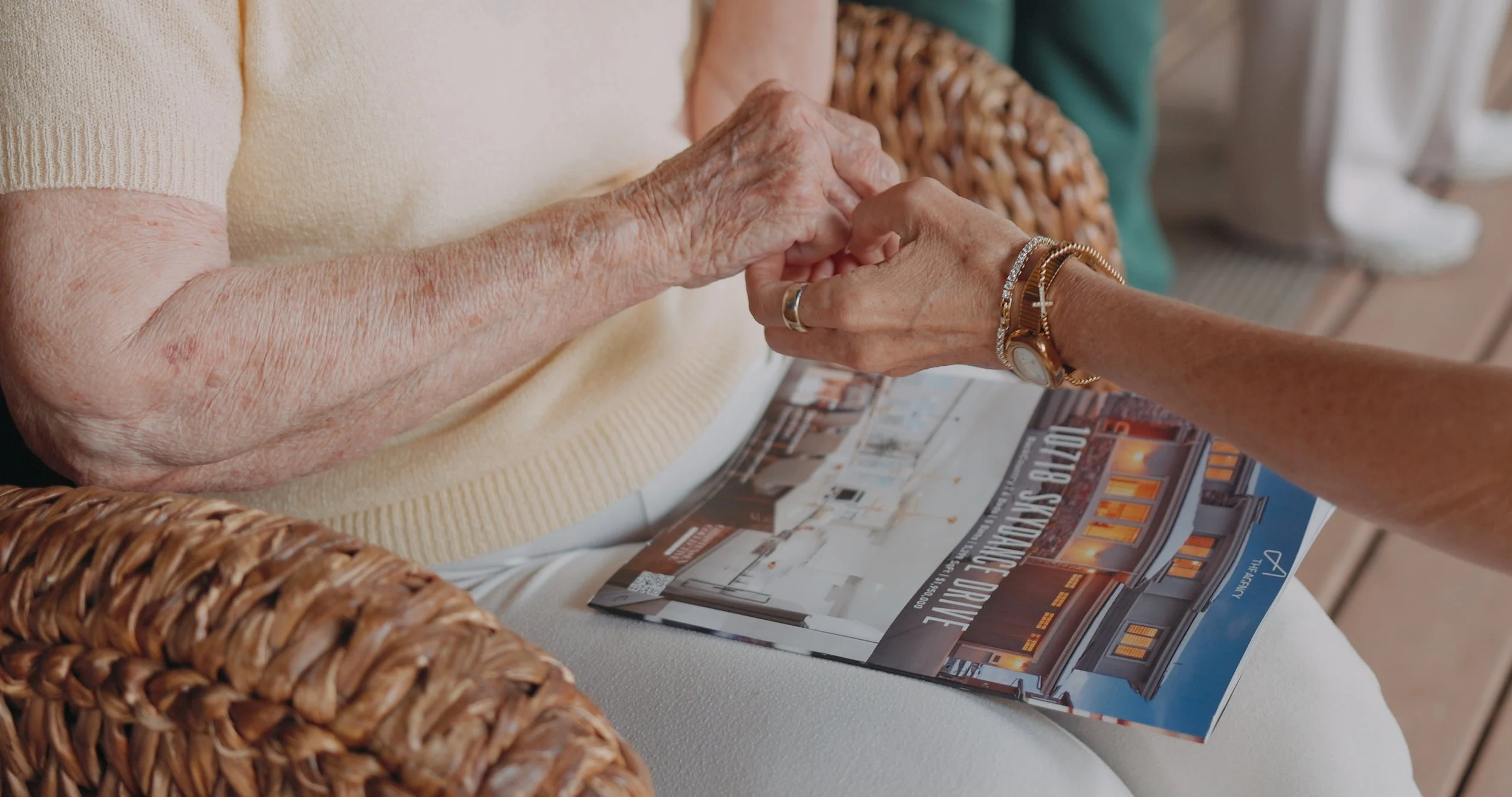 Close-up of two elderly women holding hands, one with a gold watch and bracelets, seated at a table with a real estate flyer and magazines.