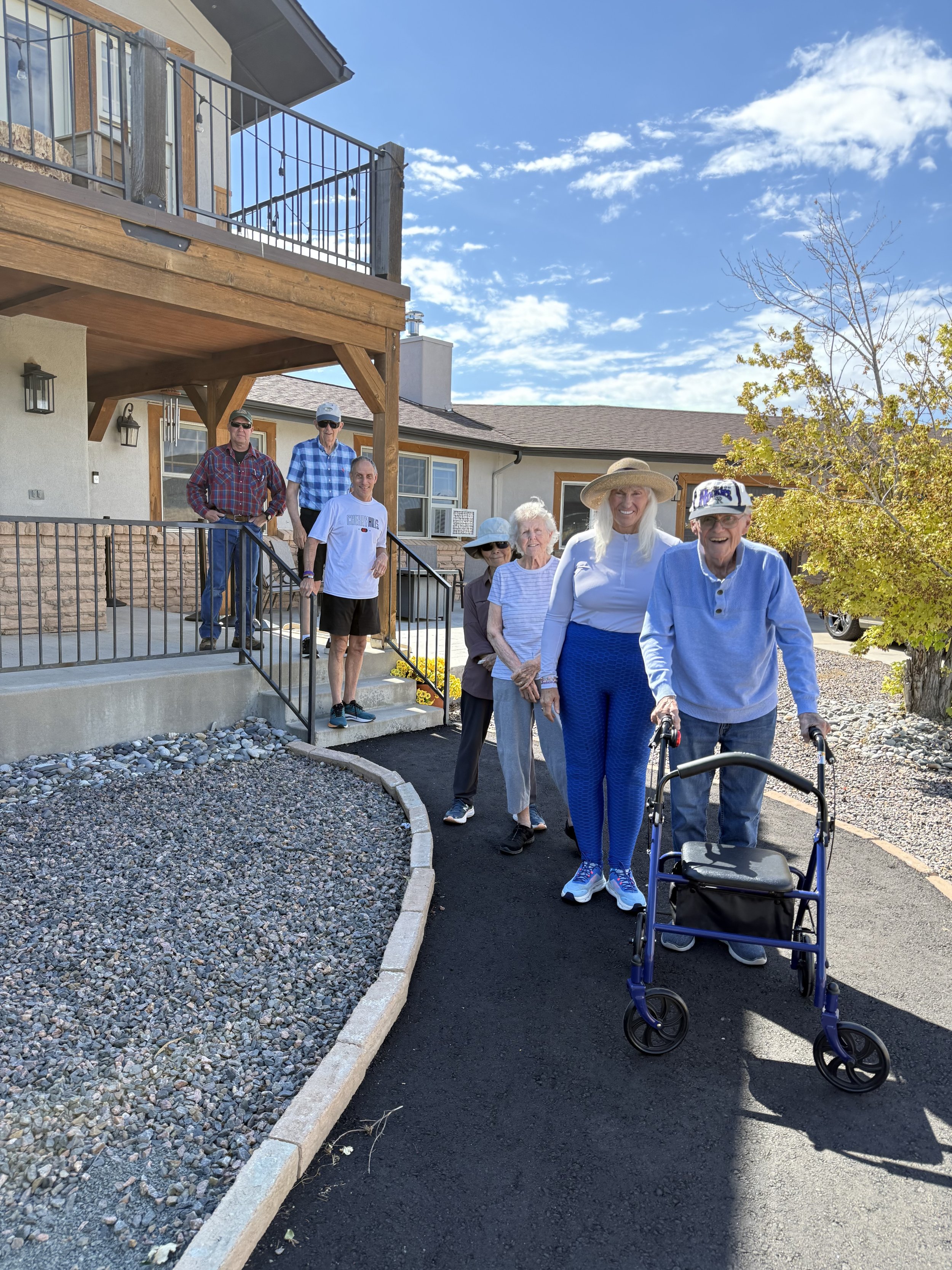 A group of elderly people walking outside a house with a new paved pathway, with some using walkers, on a sunny day.