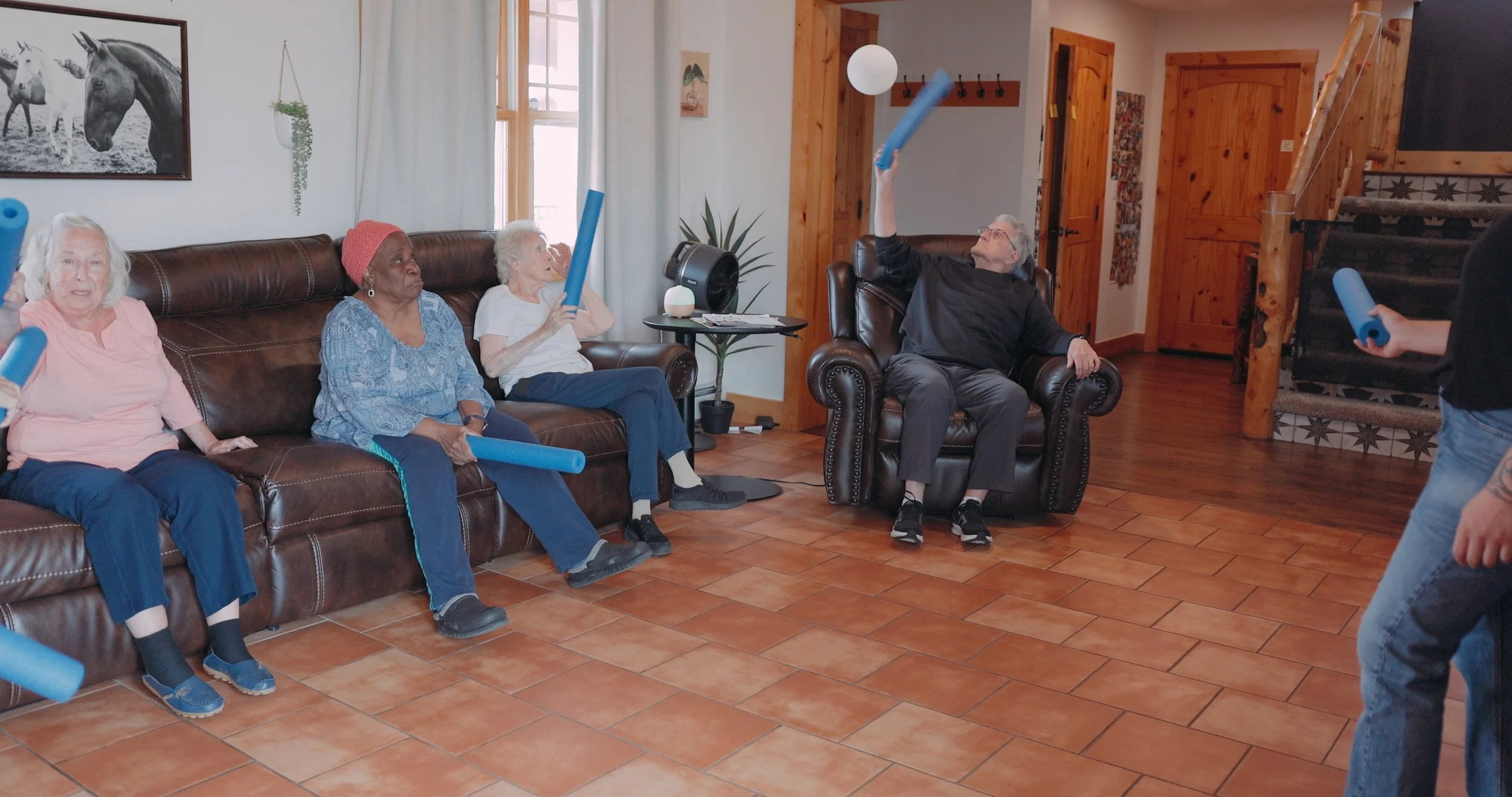 Older adults sitting on a couch and in a chair participating in a group exercise class, holding foam noodles, in a cozy living room with wooden accents.