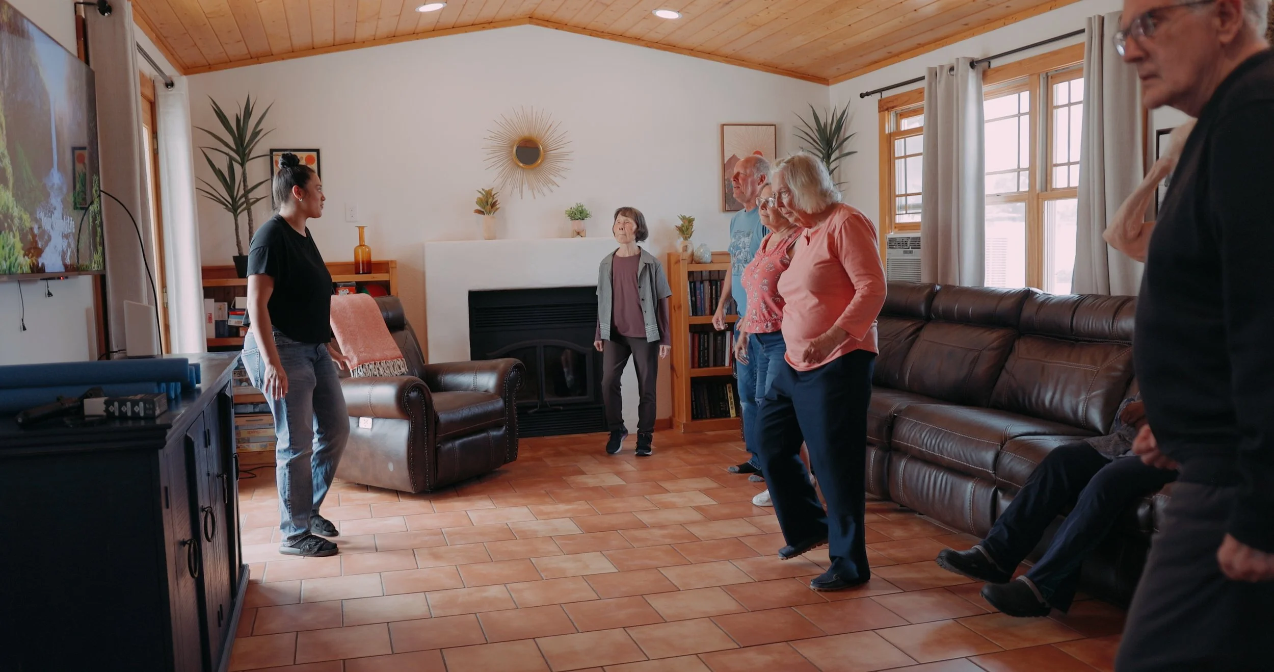 Group of elderly and middle-aged people doing a dance or exercise session in a living room, led by a young woman in black. The room has a wooden ceiling, a fireplace, bookshelves, and large windows with curtains.