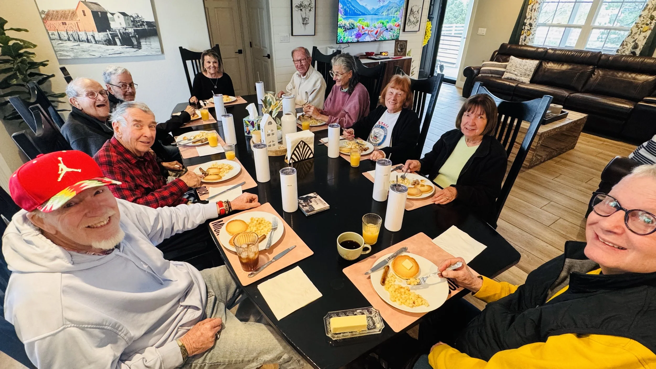 A group of elderly people sitting around a dining table, enjoying a meal together. There are plates with eggs, toast, and breakfast foods, as well as glasses of orange juice and coffee. The room has large windows, artwork on the walls, and a televisi