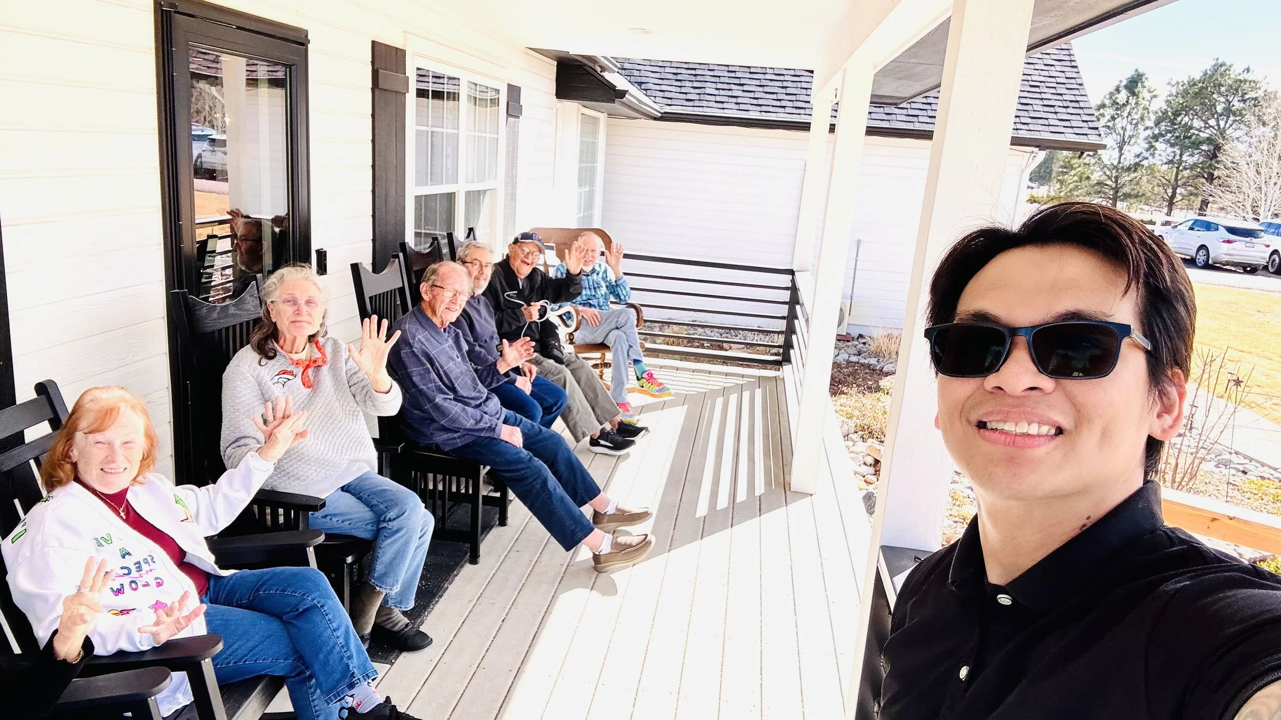 A group of elderly people sitting on a porch with a young man taking a selfie. The group is smiling and waving, and they are seated in black chairs along the house's exterior wall. The porch has white railings and a wooden floor, with a view of parke