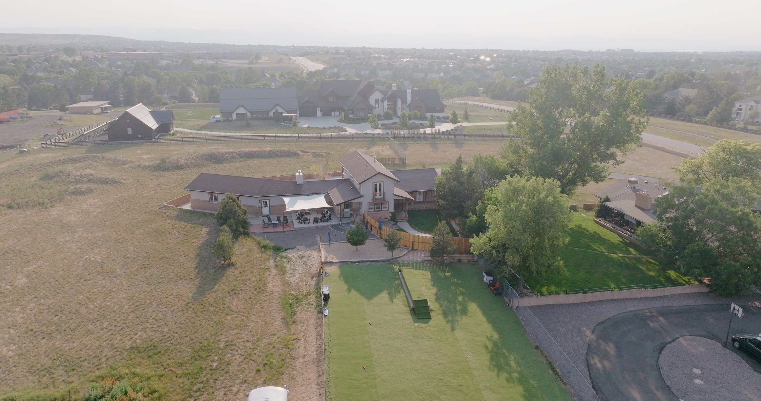 Aerial view of a residential backyard with a house, patio, lawn, tennis/basketball court, and surrounding trees.