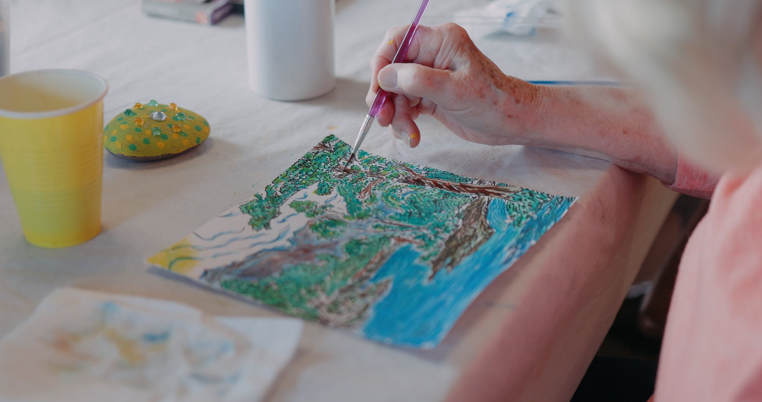 Person painting a landscape with trees, water, and mountains on paper using colored markers or pens.