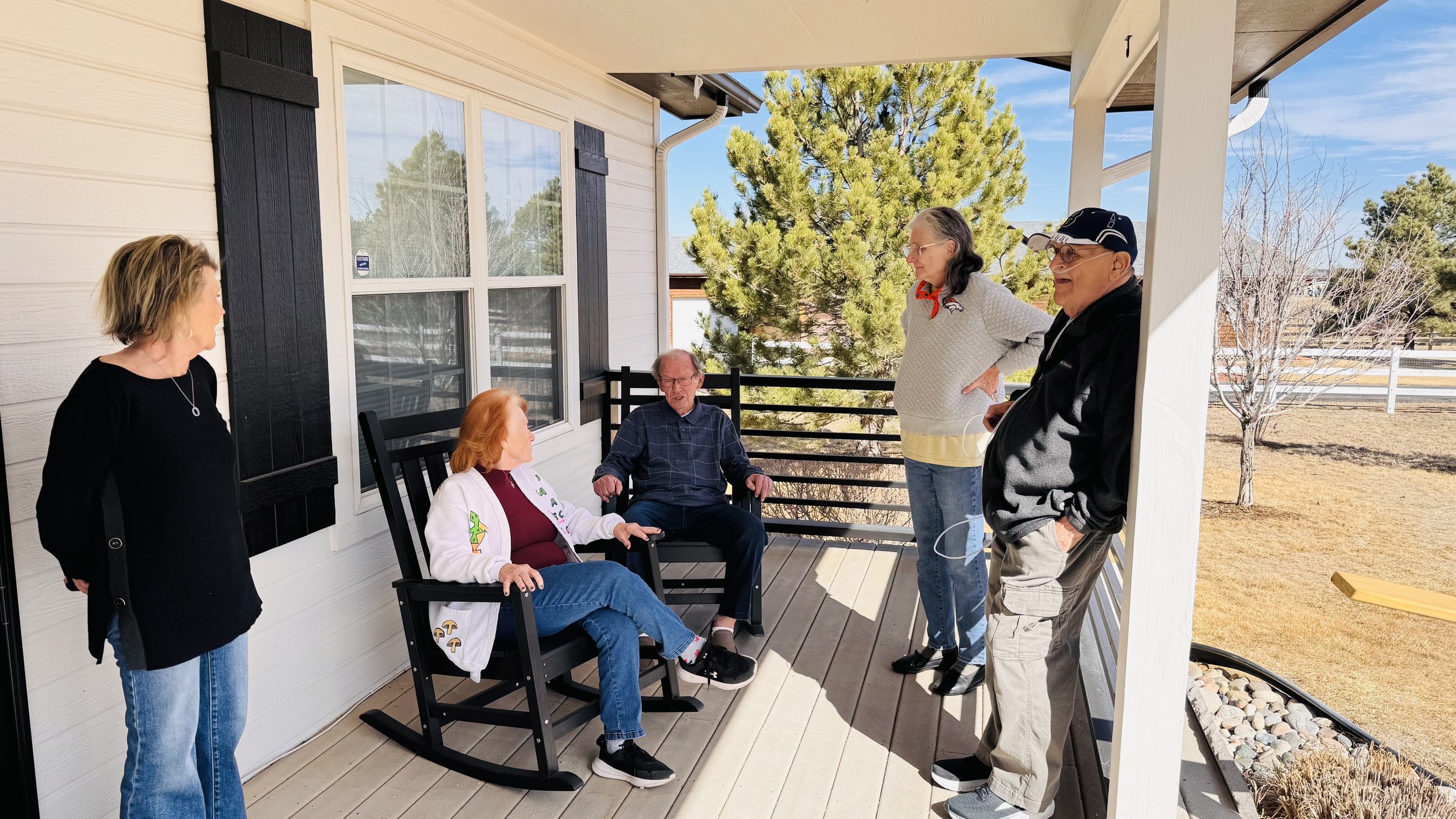 Five elderly people and one middle-aged woman conversing on a porch. Two of the elderly are seated, three others are standing. The porch is attached to a white house with black shutters. There are trees and a bright blue sky in the background.