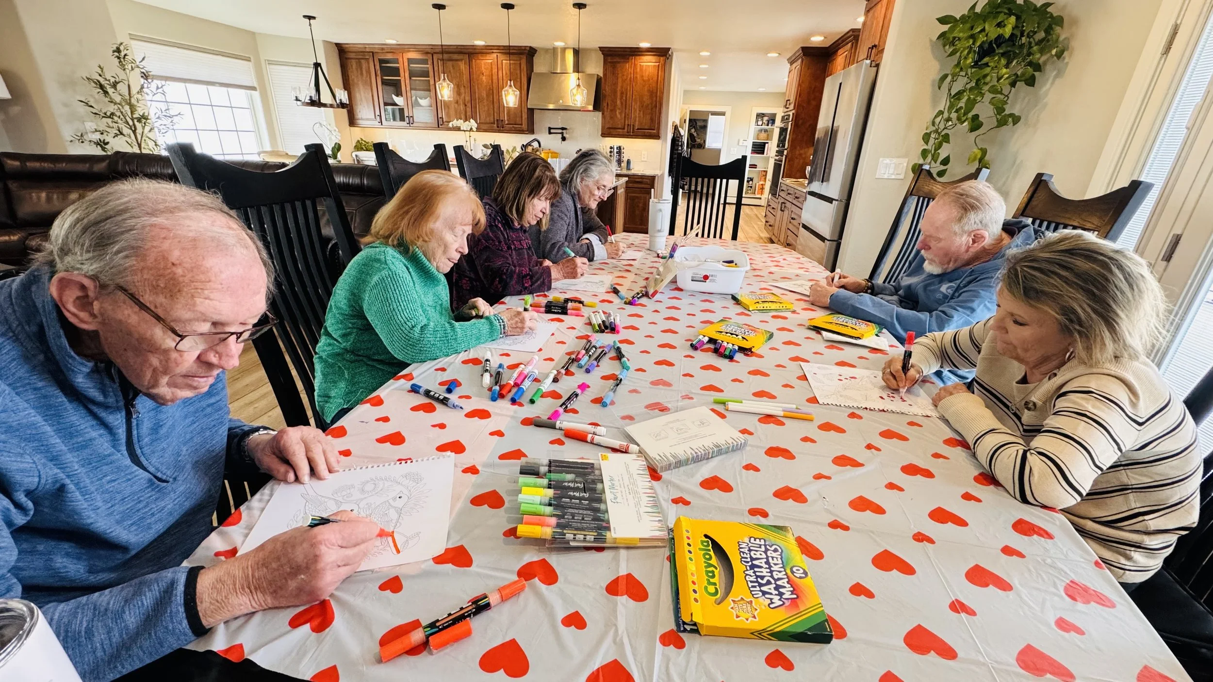 Group of elderly people sitting around a table in a kitchen, coloring with markers on sheets of paper, with Valentine-themed tablecloth and art supplies.