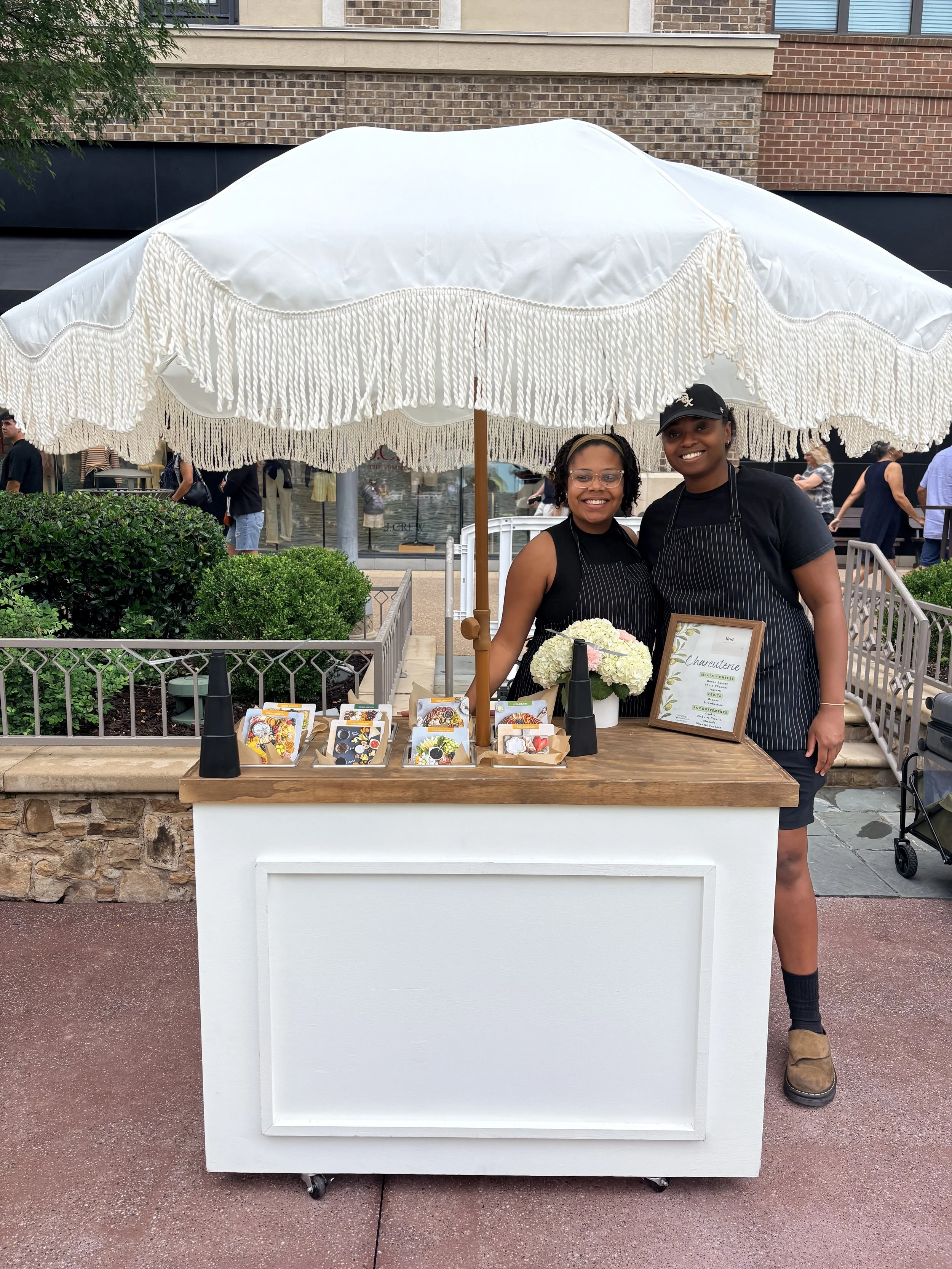 Two women smiling at a charcuterie stall under a large white patio umbrella with fringe. There are small artwork and a framed menu on the stall, and people walking in the background.
