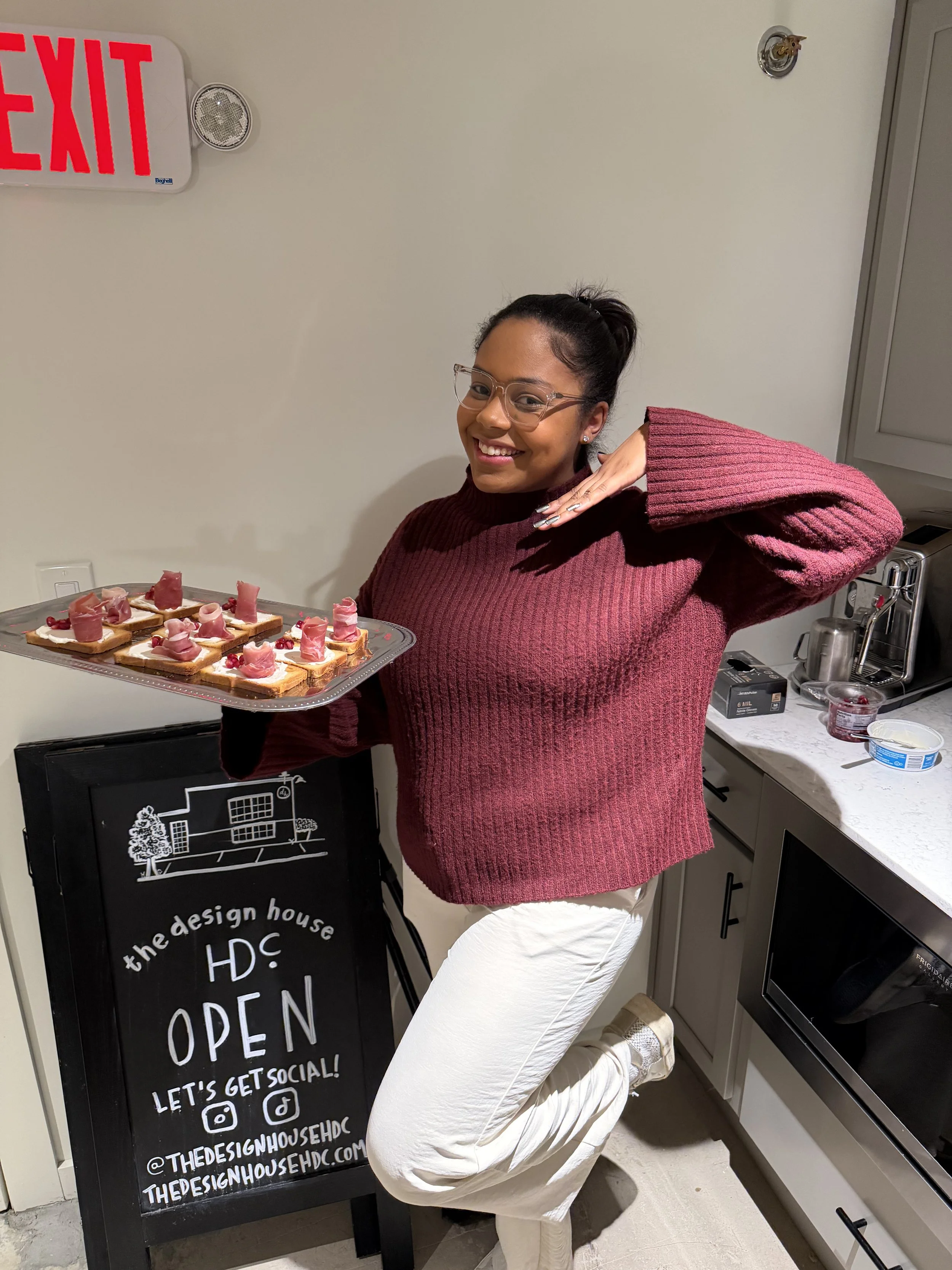 A woman with glasses, dark hair in a ponytail, wearing a burgundy sweater and white pants, is smiling and posing with her hand near her face, standing next to a tray of decorated toast or appetizers in a kitchen.