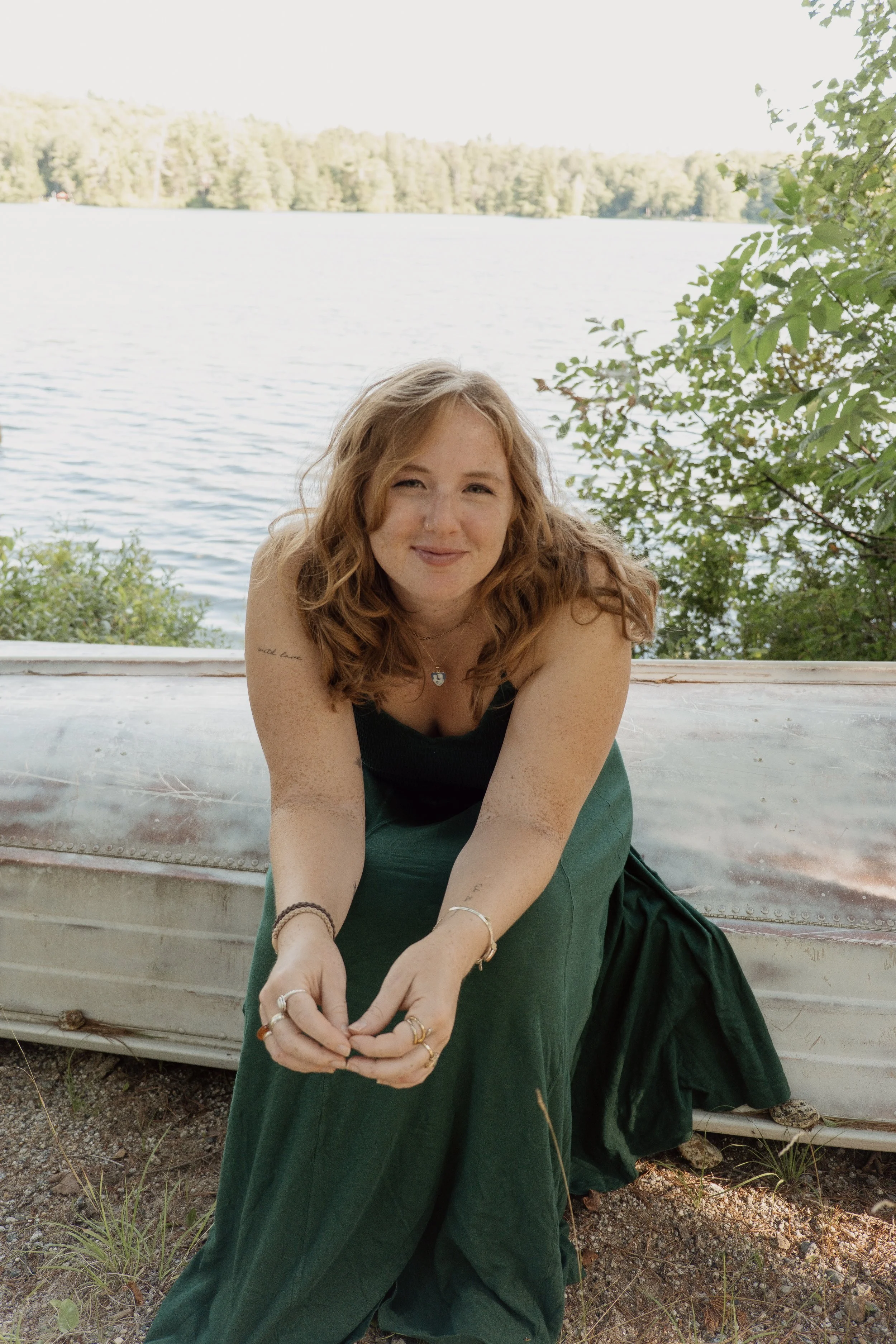 A woman with curly red hair and light skin is sitting outdoors near a lake, smiling, with trees and water in the background. She is wearing a black top and a dark green skirt, and has multiple rings and bracelets.