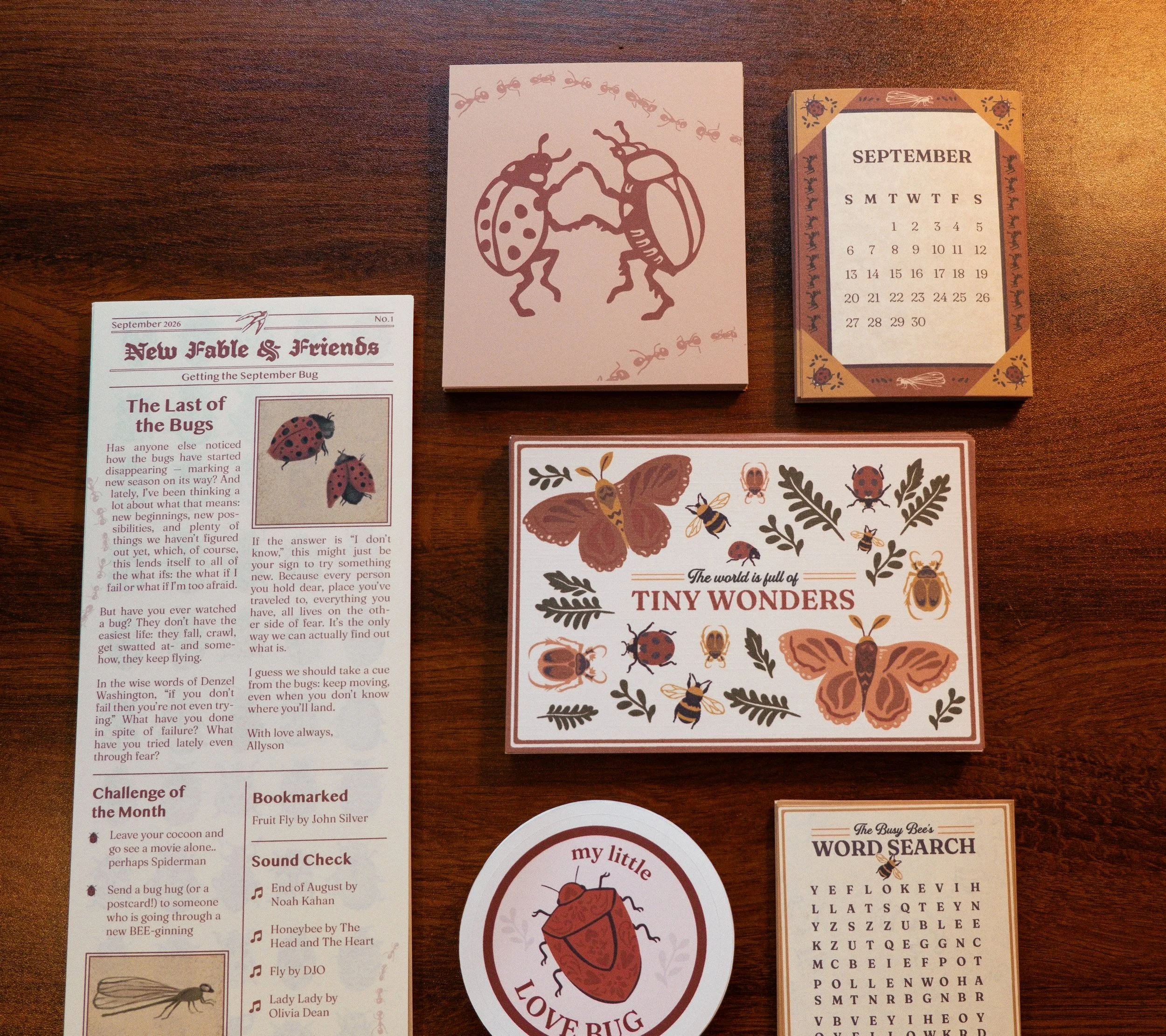Three pieces of paper on a wooden surface. The first is a newsletter page titled "New Fable & Friends" with text about bugs. The second is a pink card with a brown outline and a drawing of a ladybug, with the phrase "LOVE BUG". The third is a calendar page for September 2026 with a decorative border featuring insects and ladybugs.