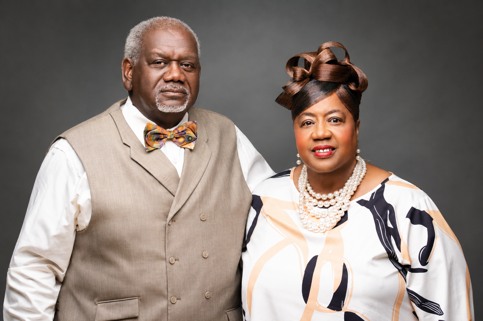 A mature African American couple dressed in stylish formal attire posing against a gray background. The man wears a beige vest, white shirt, and a colorful bow tie. The woman wears a patterned dress with a pearl necklace and earrings, and has an elaborate updo hairstyle.