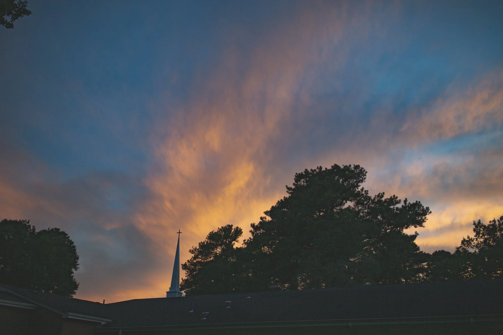 Sunset sky with clouds, trees, and a church steeple in the foreground.
