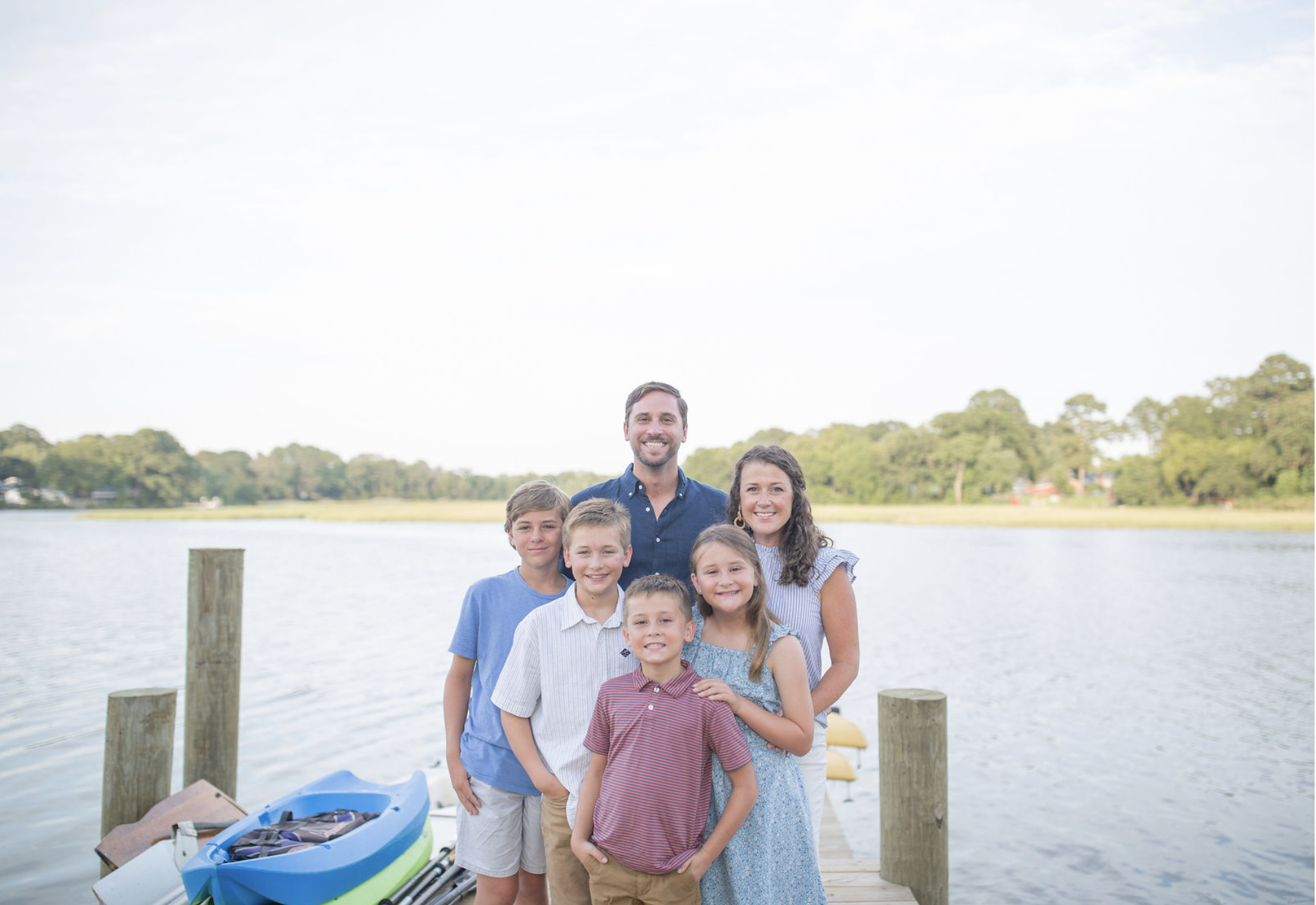 A family of eight people smiling and standing on a dock by a body of water with a green landscape in the background.