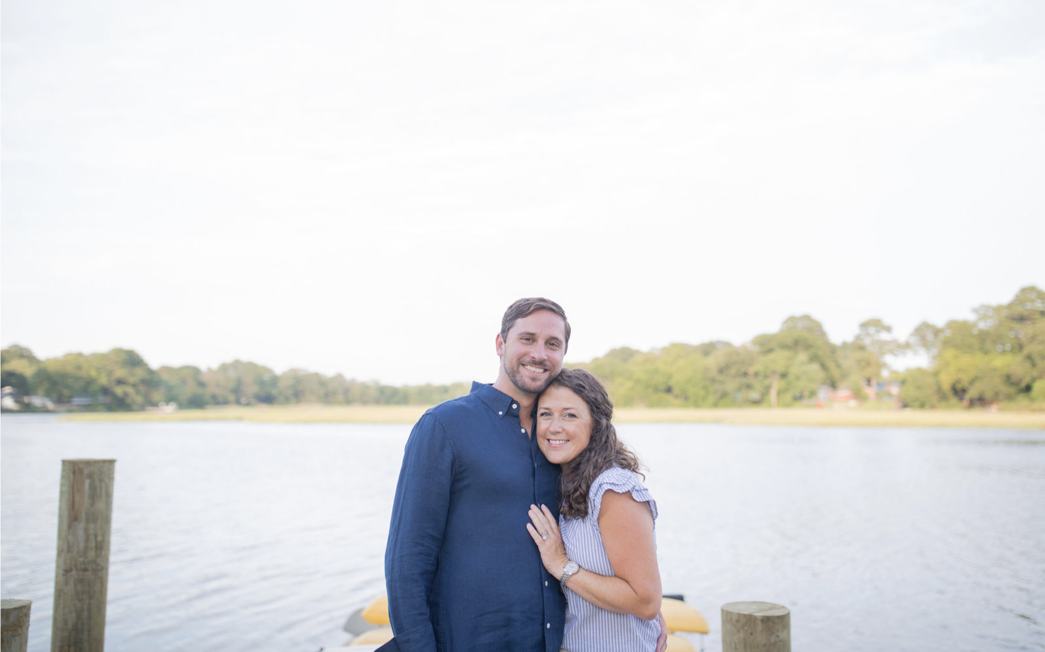 A smiling man and woman standing together outdoors near a body of water with trees in the background.