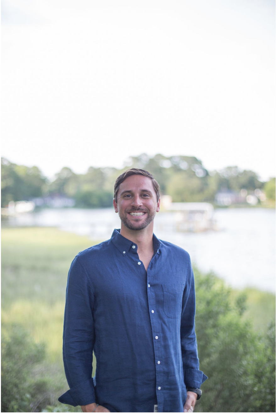 A smiling man with brown hair and a beard wearing a blue button-up shirt, standing outdoors near a body of water with trees and houses in the background.